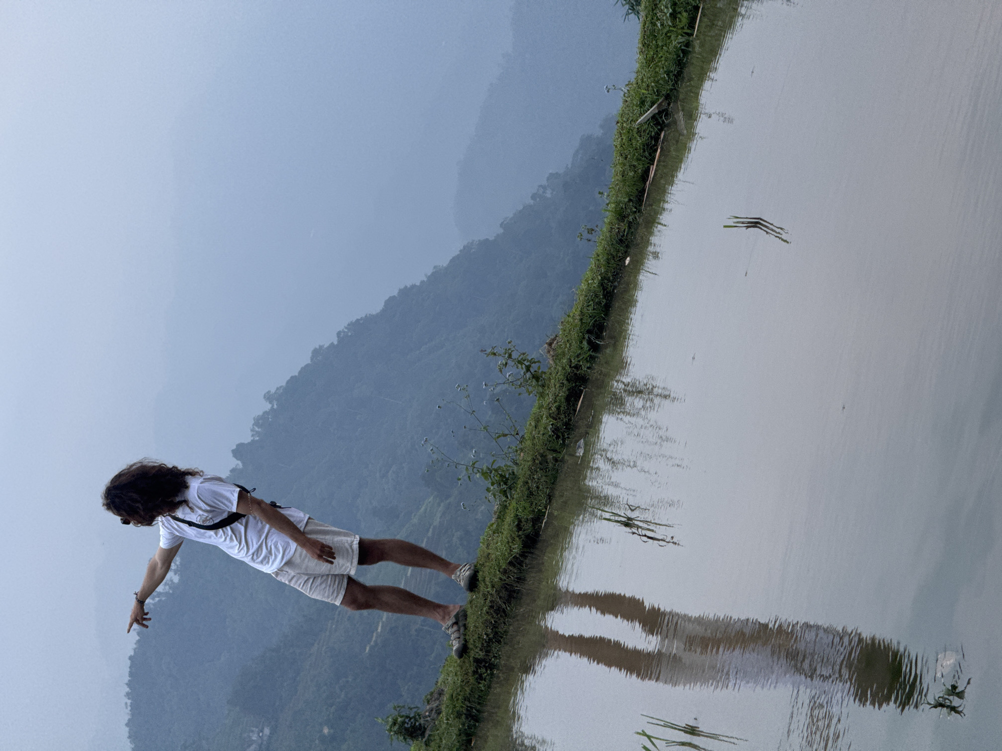 The image depicts a person striking a dynamic pose on the edge of a terraced rice field in Nậm Dịch Commune, Vietnam. The photo is originally rotated counter-clockwise by 90 degrees; when viewed correctly, it shows a person standing on a narrow, grassy embankment that separates a strip of land from a calm body of water, likely a rice paddy. In the background, layers of forested mountains recede into a soft, atmospheric haze.
The subject has long, dark, wavy hair and is wearing a white short-sleeved t-shirt, light-colored shorts, and sneakers. A dark strap, possibly from a bag, crosses their torso. They are leaning dramatically forward, with their right arm extended and finger pointing downwards towards the water's surface, as if suspended or balancing precariously. Their clear reflection is visible in the placid water to their right, mirroring their adventurous stance.
The lighting is soft and diffused, characteristic of an overcast day or early morning/late afternoon, with no harsh shadows. The distant mountains appear muted and blue-gray due to the haze, enhancing the serene and natural feel of the landscape. Small bits of vegetation are visible floating on the water's surface and along the grassy edge of the embankment. There is no discernible text present in the image.