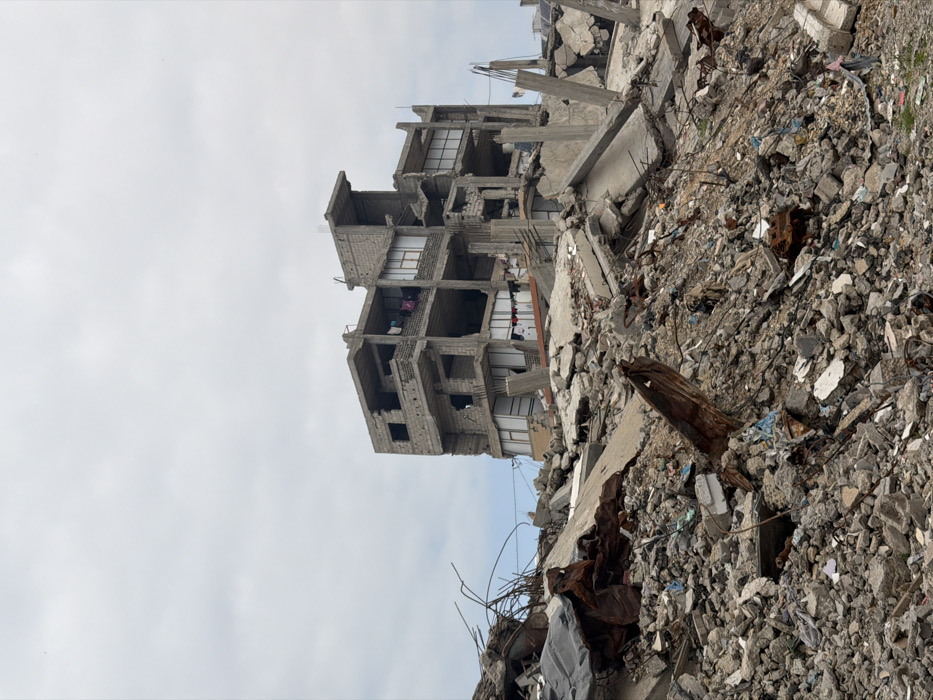A destroyed building stands amidst rubble under a cloudy sky. The multi-story structure is heavily damaged, revealing exposed rooms. Debris consisting of concrete, metal, and various materials piles up around the building's base, reaching a significant height. The scene conveys devastation and destruction.