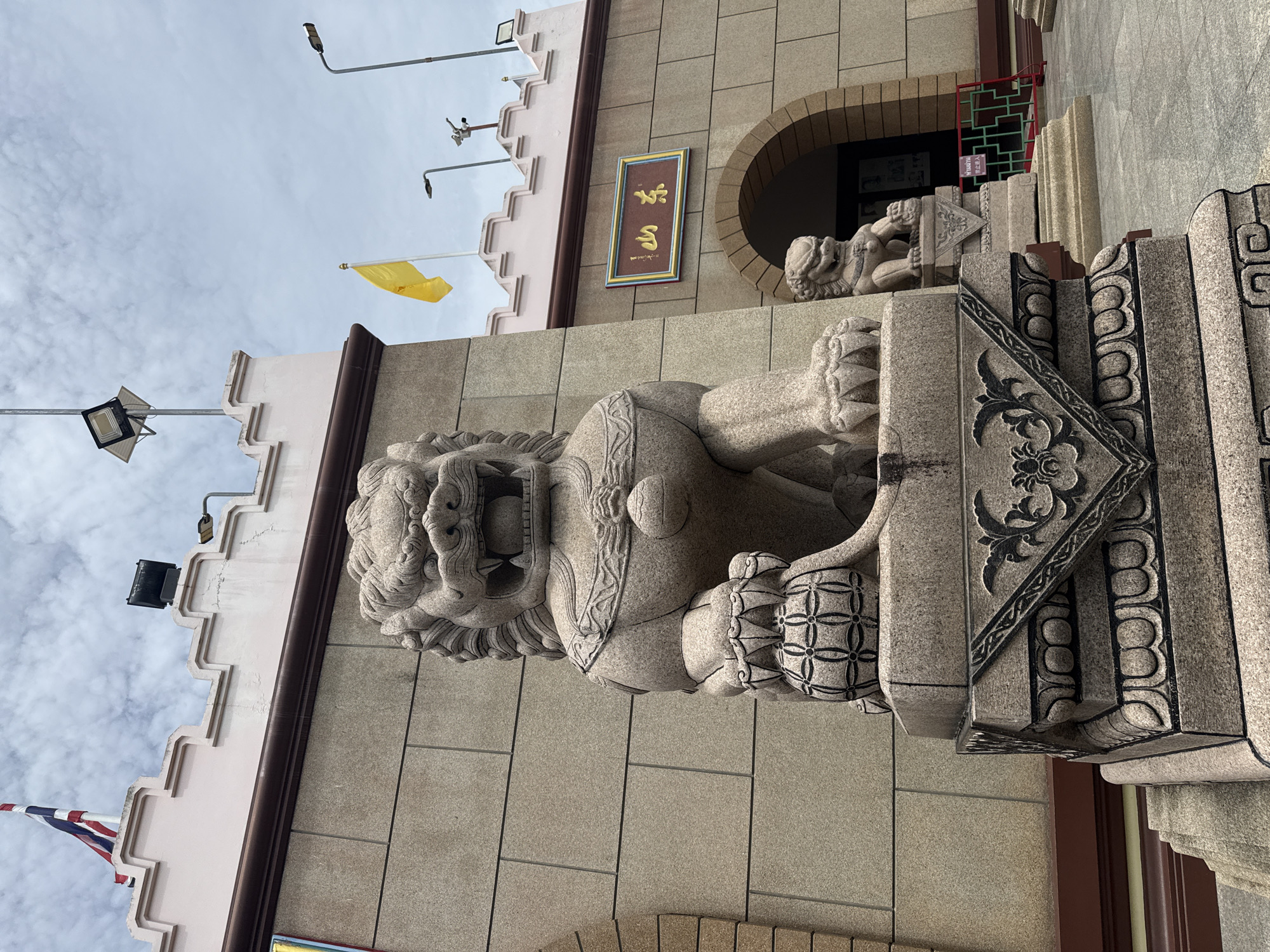In a medium shot, two stone lion statues stand guard in front of a building with a crenellated pink parapet. The foreground statue, a large, intricately carved lion with a fierce expression, is positioned on a stone pedestal adorned with black floral and geometric patterns. The lion is seated, with its front paws resting on a decorative spherical object. To the right and further back, a smaller, similar lion statue sits on a matching pedestal.
The building behind the statues has a light beige, stone-block facade. Above the arched entrance, a rectangular sign with gold lettering on a blue background is mounted. A yellow flag flies from a pole on the roofline. The sky is overcast with scattered clouds. The overall scene appears to be an outdoor location in the city of มาบฟักทอง, Thailand, likely a temple or cultural site given the architectural style and statues.