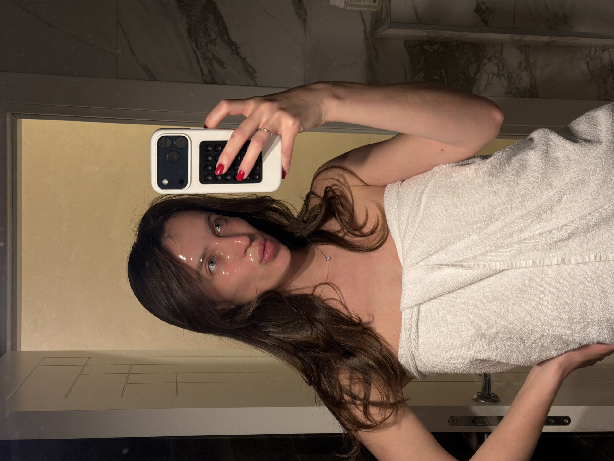 A woman wearing a white towel is in a bathroom in Budva, Montenegro, taking a selfie in a mirror. The bathroom features a marble countertop and a black tiled floor. She has a clear face mask on, with water droplets visible. Her hair is long and brown, and she is wearing a silver necklace and a ring. Her fingernails are painted red. She is holding a white smartphone with a black and white phone case.