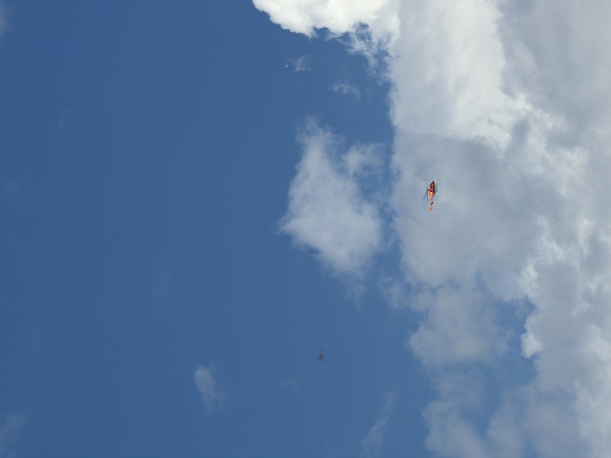The image shows a bright blue sky with scattered white clouds. A small, orange helicopter is visible in the lower center of the frame, flying in the distance. The helicopter has two main rotors and a tail rotor. The overall impression is one of a clear day with a helicopter in flight. No specific landmarks of Saint Petersburg are identifiable in this image.
