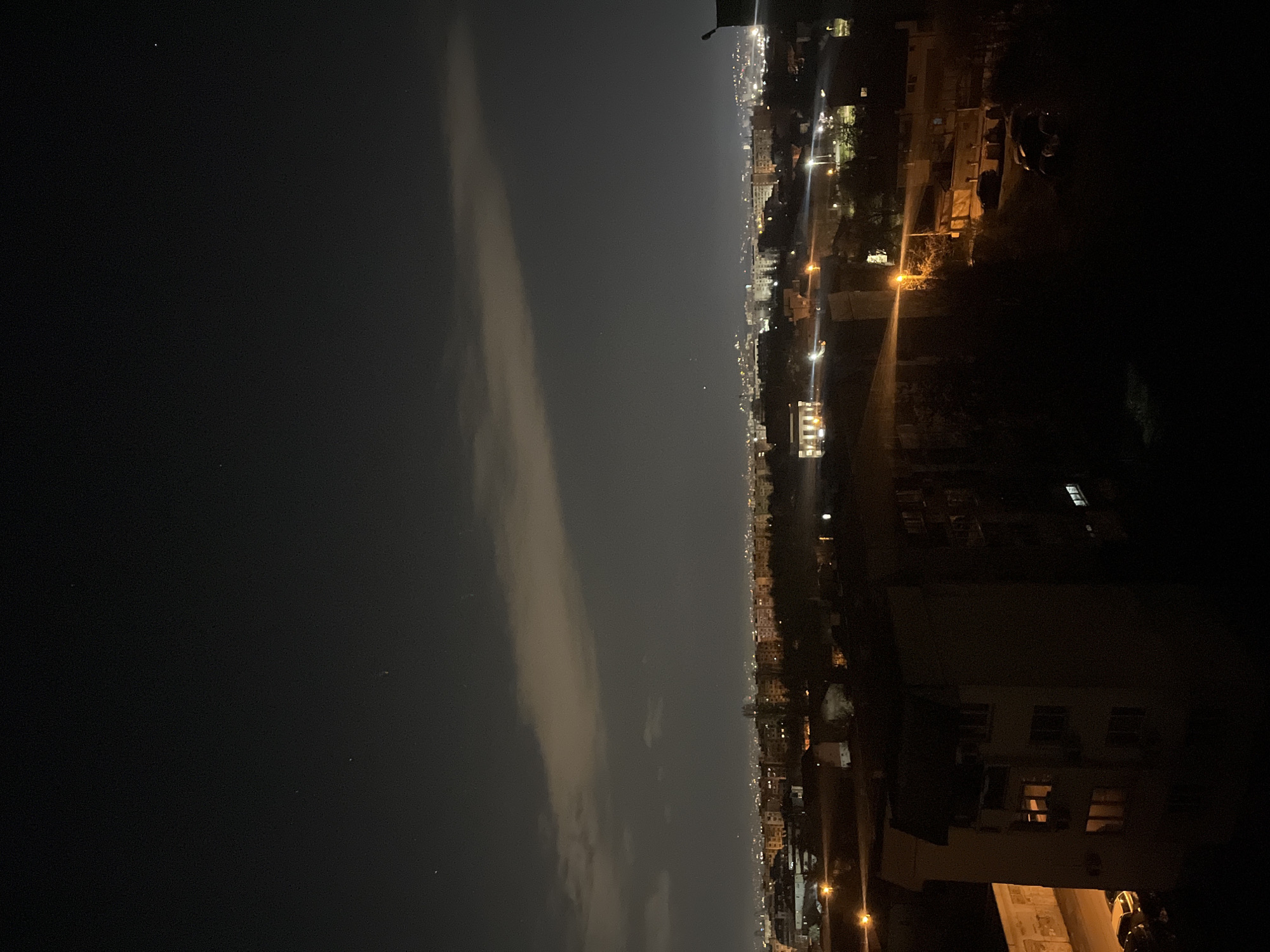 The image shows a night view of Sofia, Bulgaria. The sky is dark with some clouds. In the distance, city lights illuminate the skyline. Some residential buildings are visible in the foreground on the right side of the image. The scene is generally dark.