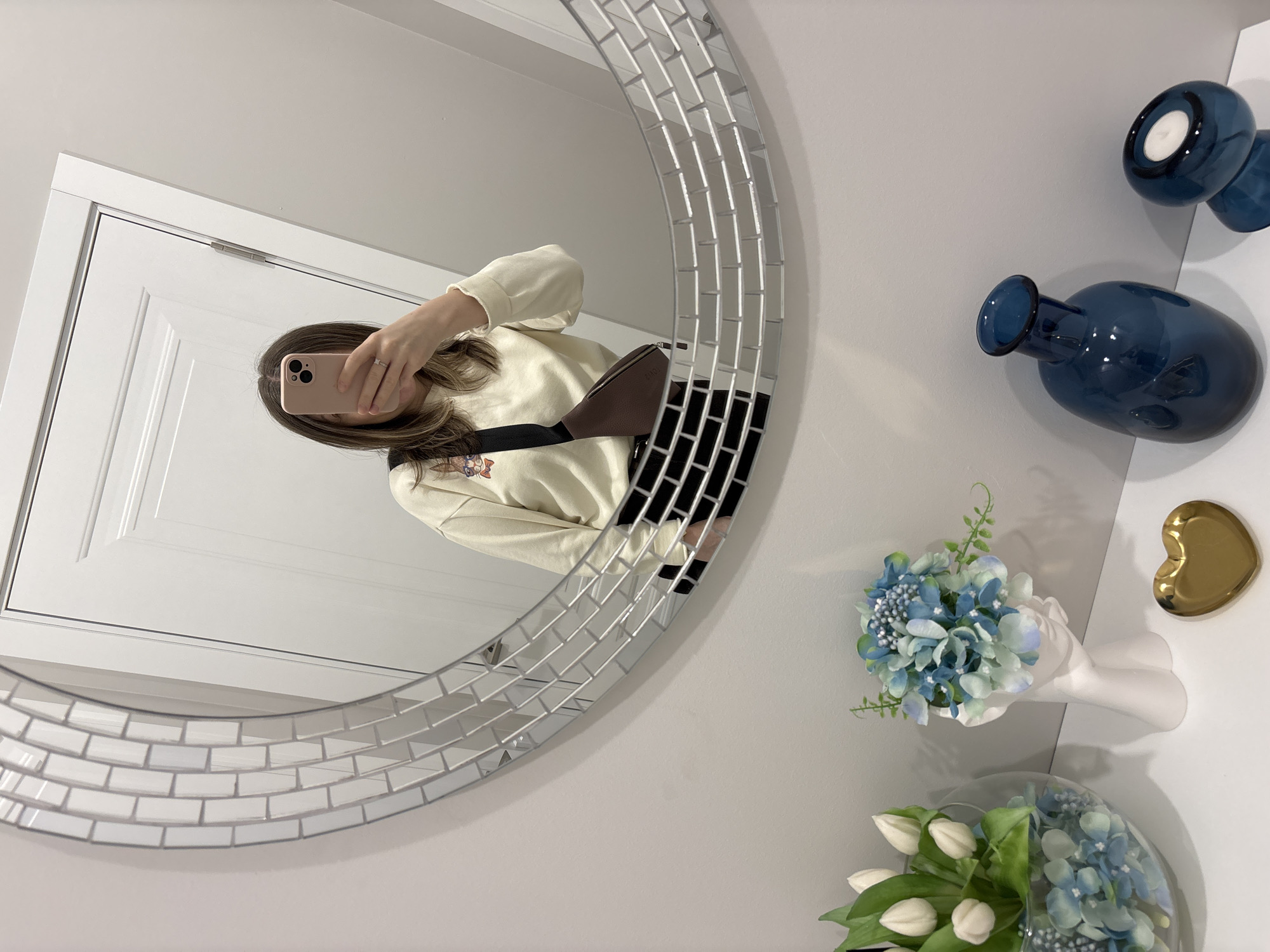 A selfie is captured in a round mirror with a decorative, brick-like frame. The reflected person, holding a phone, is wearing a beige sweater and a brown bag across their body. The background visible in the mirror is a white door with recessed panels. To the right of the mirror, on a light-colored wall, are two small blue glass vases, a gold heart-shaped dish, and two floral arrangements. The flowers appear to be hydrangeas and tulips in a white vase. The setting appears to be a bathroom or dressing area. There is no information to indicate the location is in Saint Petersburg, Russia.