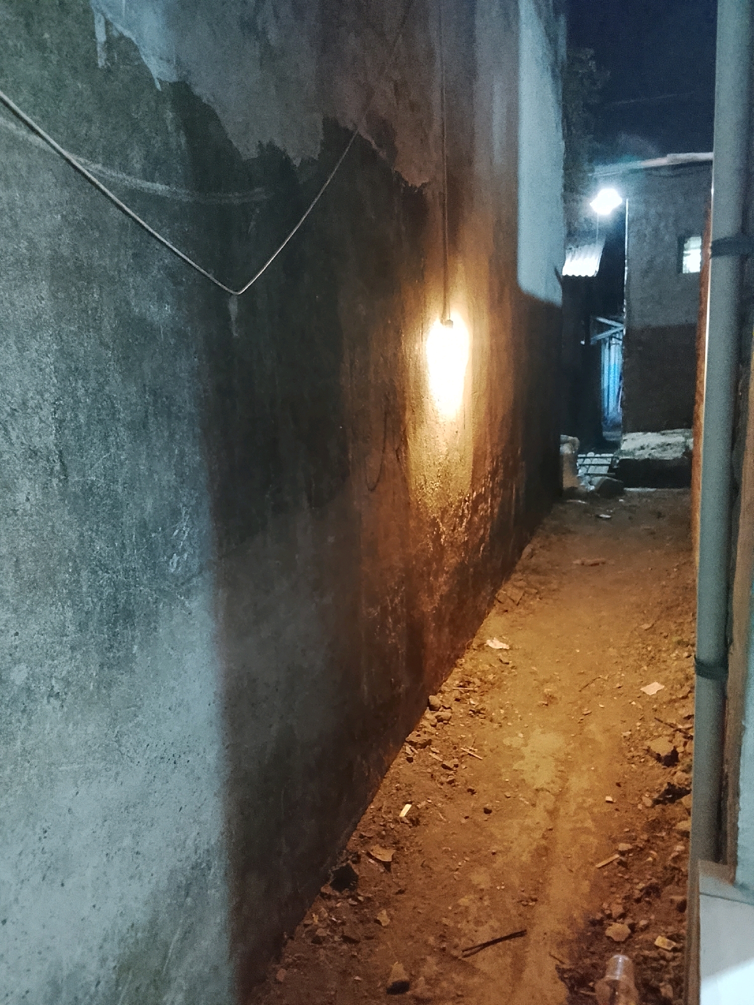 The image depicts a narrow, dimly lit alleyway in Ujungrusi, Indonesia, at night. The ground is uneven and covered with dirt, small rocks, and scattered debris. On the left, a rough, textured wall dominates the scene, with a single bare light bulb hanging from a wire, casting a warm, yellowish glow. The light illuminates a portion of the wall and the ground directly beneath it, creating a stark contrast with the surrounding darkness.
To the right, another wall is visible, also appearing to be made of concrete or plaster. Further down the alley, a structure with a corrugated roof can be seen, and in the background, there are hints of other buildings and a small window with light emanating from it. A vertical pipe runs along the right edge of the frame, partially obscuring the view of the wall behind it. The overall atmosphere is one of urban neglect and the quiet solitude of a deserted alley at night. There are no people or animals visible in the image.