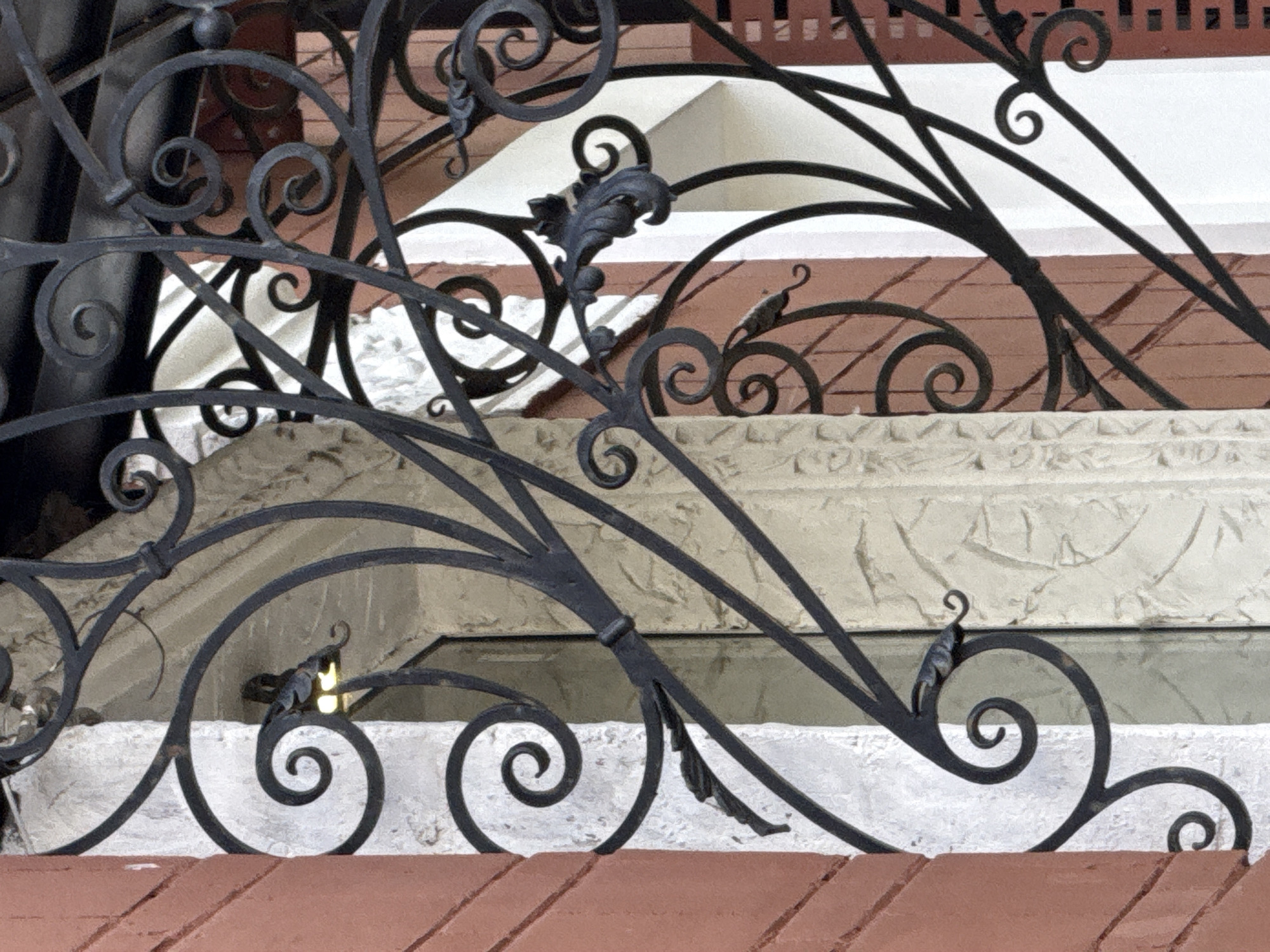 The image shows a close-up of ornate black wrought ironwork in front of a building. The ironwork features elaborate scrollwork, leaf motifs, and curled decorative elements. Behind the ironwork, there is a textured light-colored stone or plaster surface, a reddish-brown brick wall, and parts of a window or doorway. A dim light source is visible through the ironwork. The style of the ironwork suggests it is an architectural detail, possibly a balcony railing, gate, or window grille. The building facade is characterized by a combination of materials, including the brickwork and the decorative stone surround. The overall impression is one of historical or traditional architectural design.