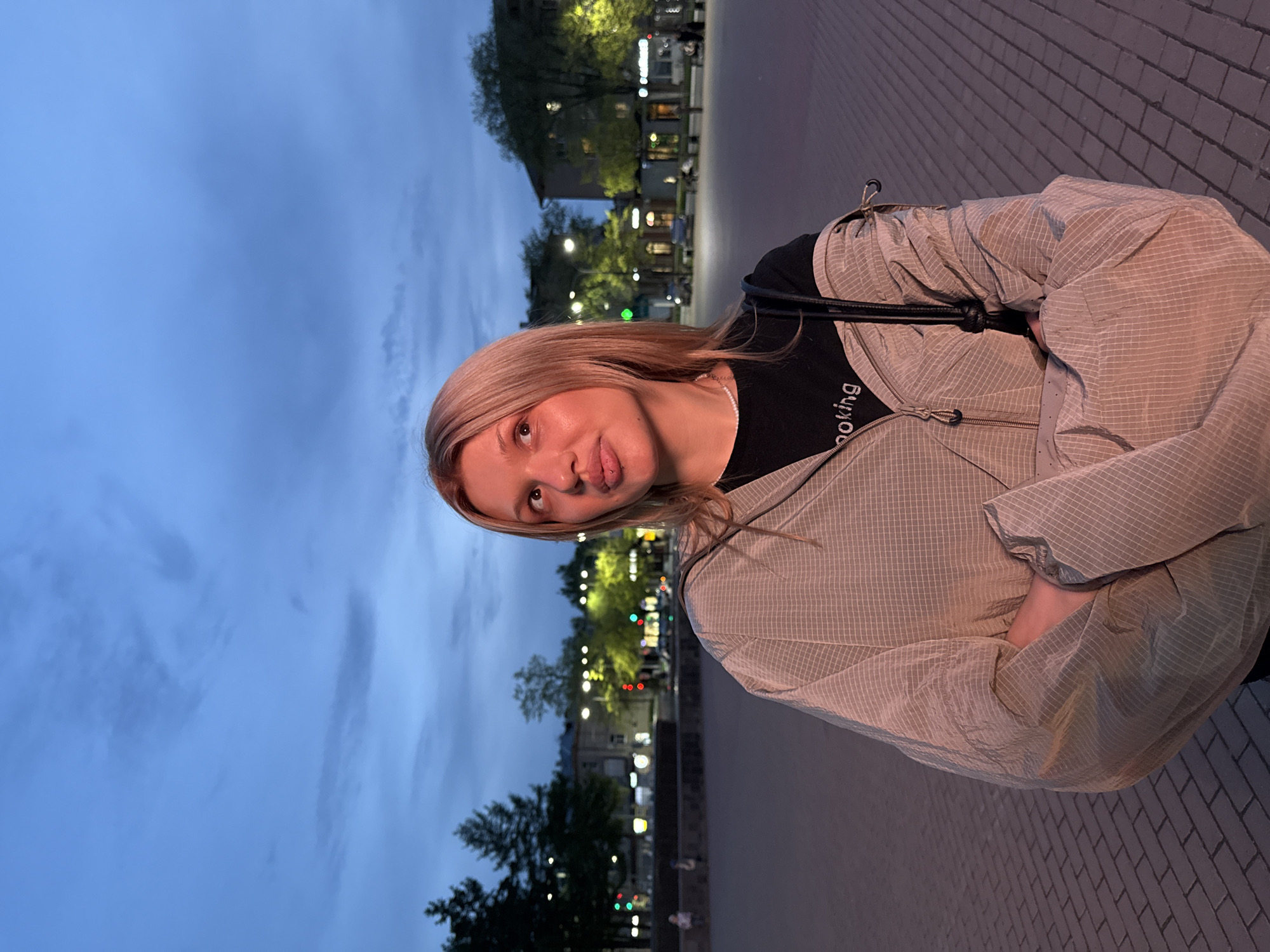 A woman with blond hair stands in a city square in Chaikovsky, Russia, at dusk. She is wearing a black shirt with white lettering and a beige jacket. She is standing on a brick-paved area with trees and buildings visible in the background. Streetlights are illuminated.
