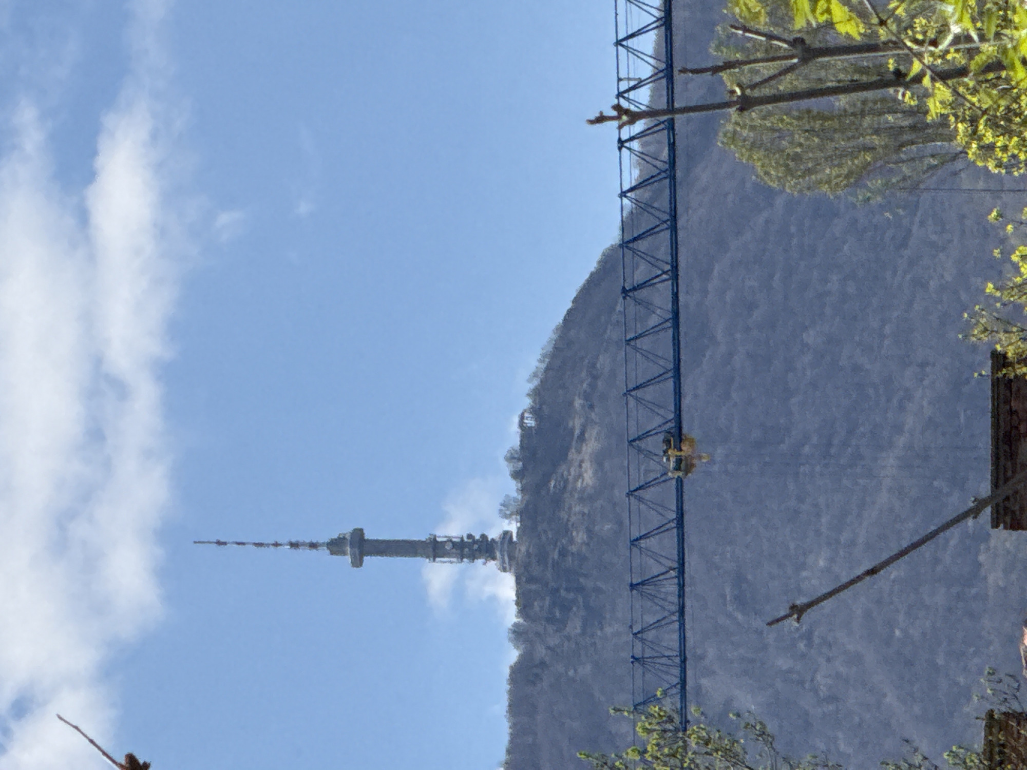The image shows a view of the Kopitoto TV tower in Sofia, Bulgaria. The tower is located on a hill, with a blue, metal suspension bridge to its right. The sky is blue with some white clouds. Some foliage is visible on the right side of the image.