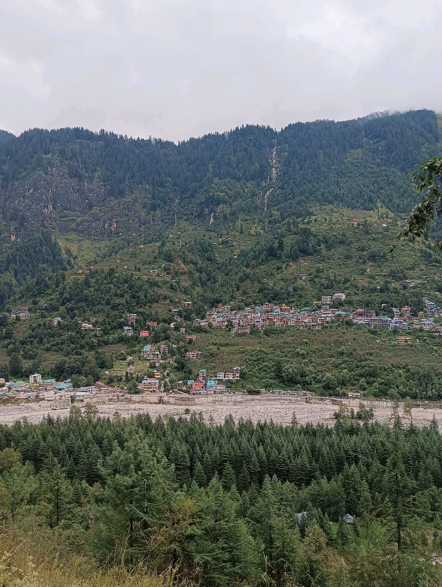 The image depicts a wide shot of the mountainous region surrounding Vashisht, India. In the foreground, a dense forest of pine trees covers the lower slopes. Beyond the forest, a wide, rocky riverbed is visible, with a small settlement of buildings situated along its banks and extending up the hillside. These buildings are of various sizes and colors, with many featuring multi-story structures. Further up the mountain, more buildings are scattered amongst the trees, indicating a higher density of habitation. The background is dominated by a steep, forested mountain range under a cloudy sky. Waterfalls are faintly visible cascading down the slopes of the mountain. The overall scene conveys a sense of a remote, mountainous town nestled within a lush, green landscape.
