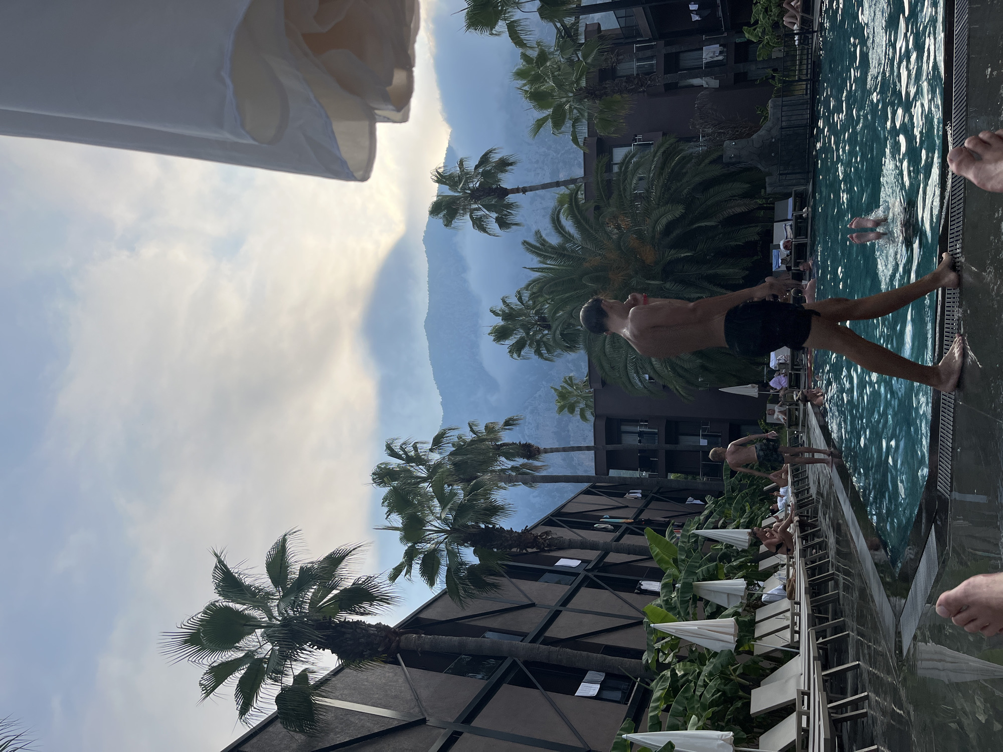 The image depicts a resort setting with a swimming pool and palm trees, under a cloudy sky with mountains in the background. In the foreground, a young person walks along the edge of the pool, wearing black swim trunks. Another person is partially submerged in the pool. Several lounge chairs are arranged by the pool, some occupied by people. The architecture of the resort buildings is visible, with dark brown facades and geometric patterns. The overall atmosphere suggests a vacation or leisure activity.