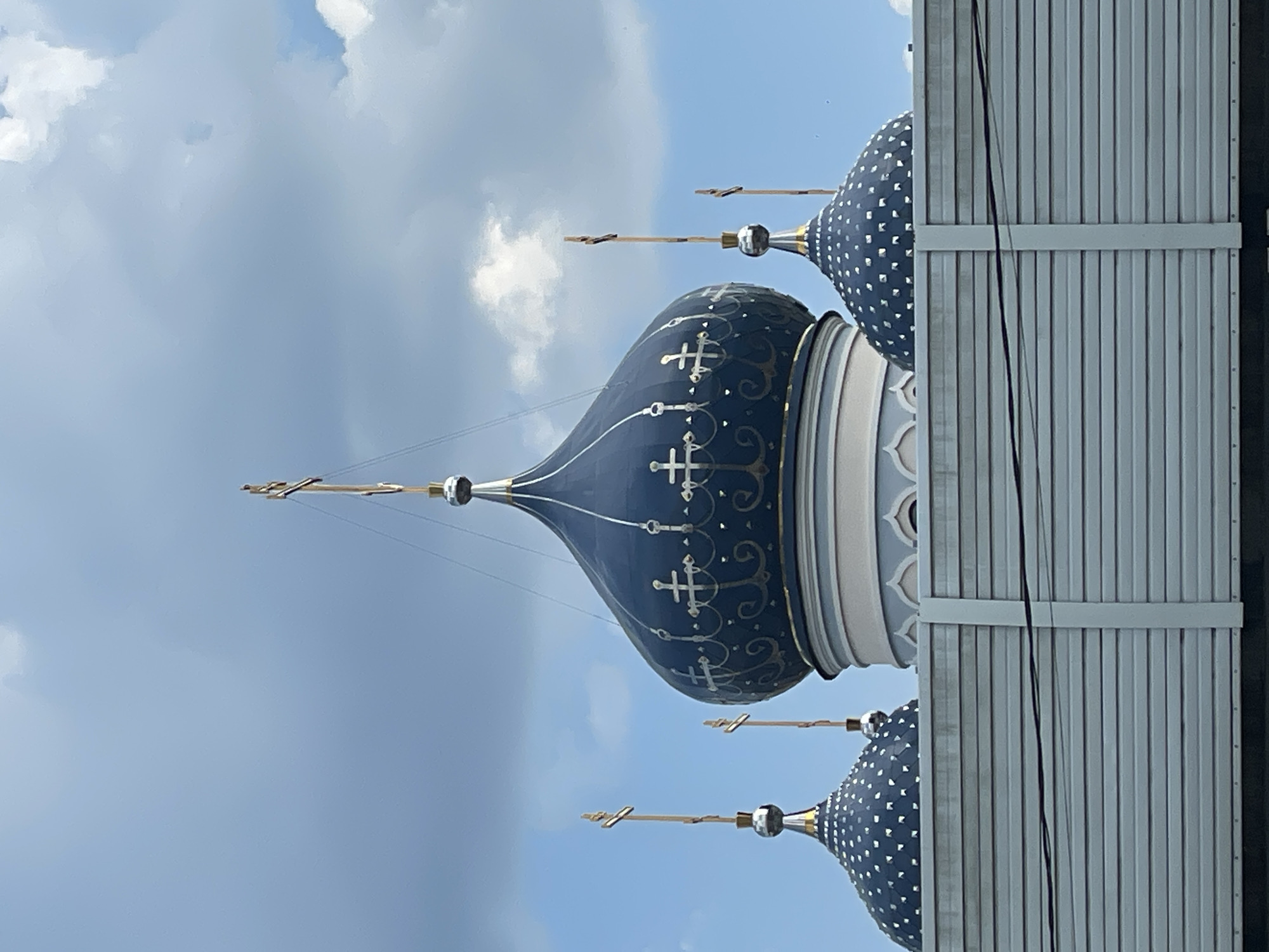 The image shows the domes of an Orthodox church against a cloudy blue sky, partially obscured by a grey wall. The larger dome is blue with gold crosses and decorative swirls, while the smaller dome is blue with silver dots. It is located in Moscow, Russia.