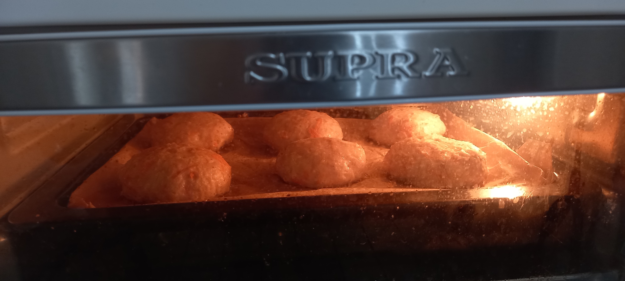 A tray of six evenly spaced, raw, light-colored balls, likely meatballs or dough, is baking inside a "SUPRA" brand oven. The tray is lined with parchment paper, and the oven light is on. The setting appears to be indoors. The location is unknown.