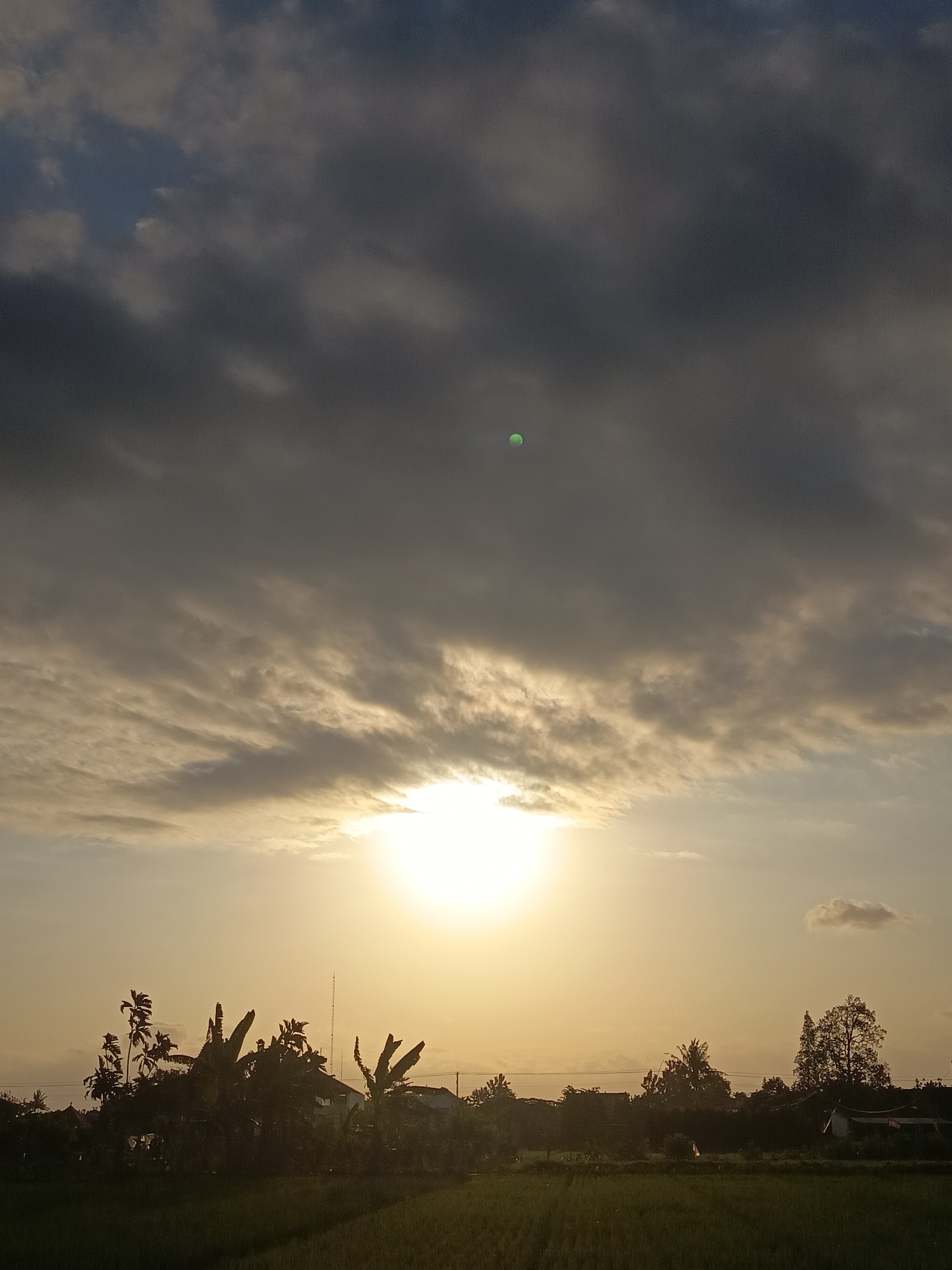 The image depicts a sunset in Sidokarto, Indonesia. The sky is filled with dramatic, dark clouds, with the bright sun peeking through the lower half. A faint, small green orb is visible in the upper portion of the sky. Silhouetted against the sunset are lush green rice fields in the foreground. Behind the fields, a line of trees and vegetation forms a dark silhouette, with some structures, likely houses, partially visible amongst them. A tall, thin antenna is also present, standing out against the twilight sky. The overall atmosphere is serene and natural.