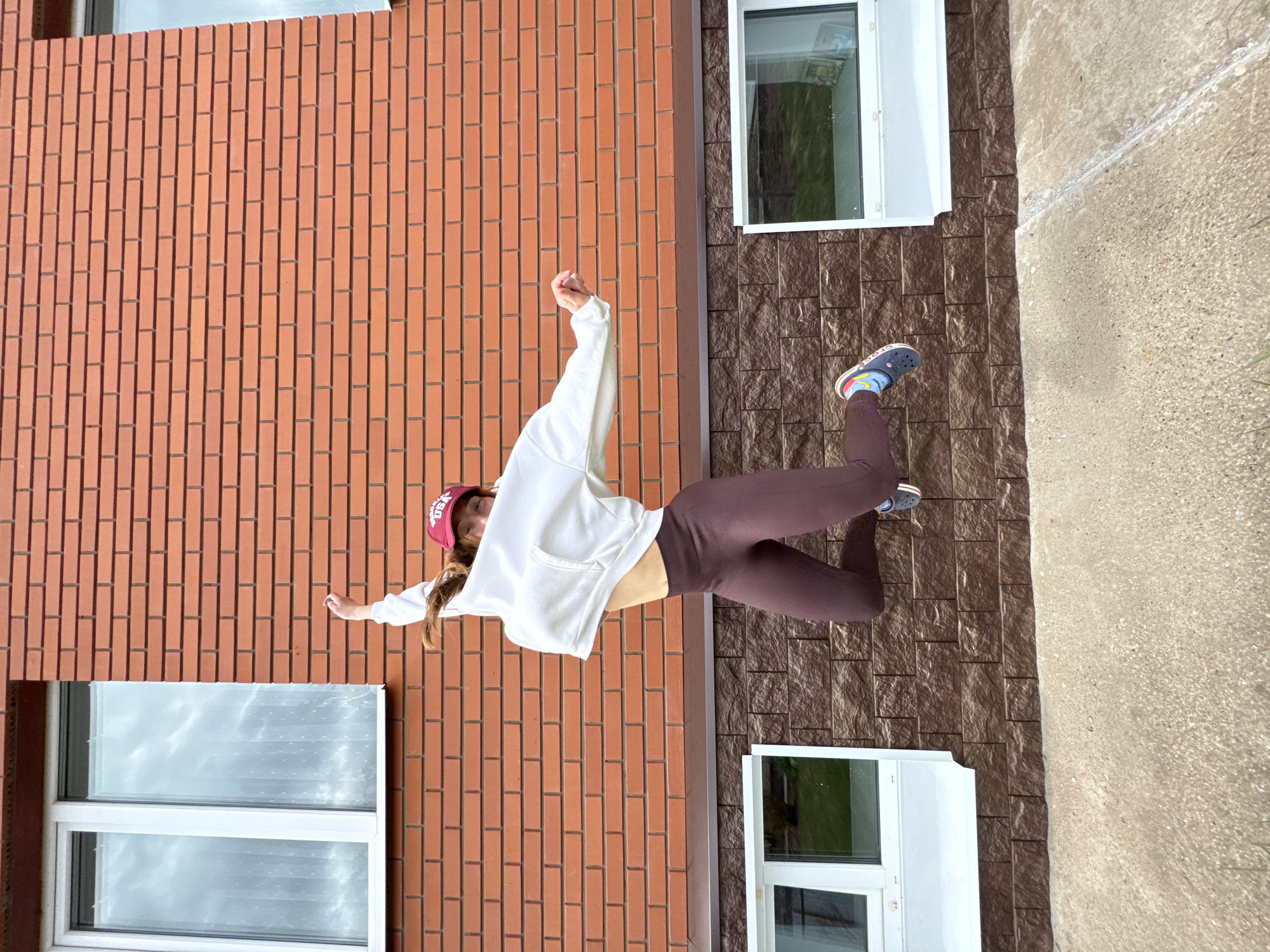 A vertical shot depicts a young woman standing against a building in Прикамский, Russia. The building features red brick on the left and textured brown material on the right. A window with white trim is located on each side of the woman. The woman is wearing a white hoodie, maroon leggings, and a red baseball cap. She has her arms outstretched. The ground is a light gray concrete.