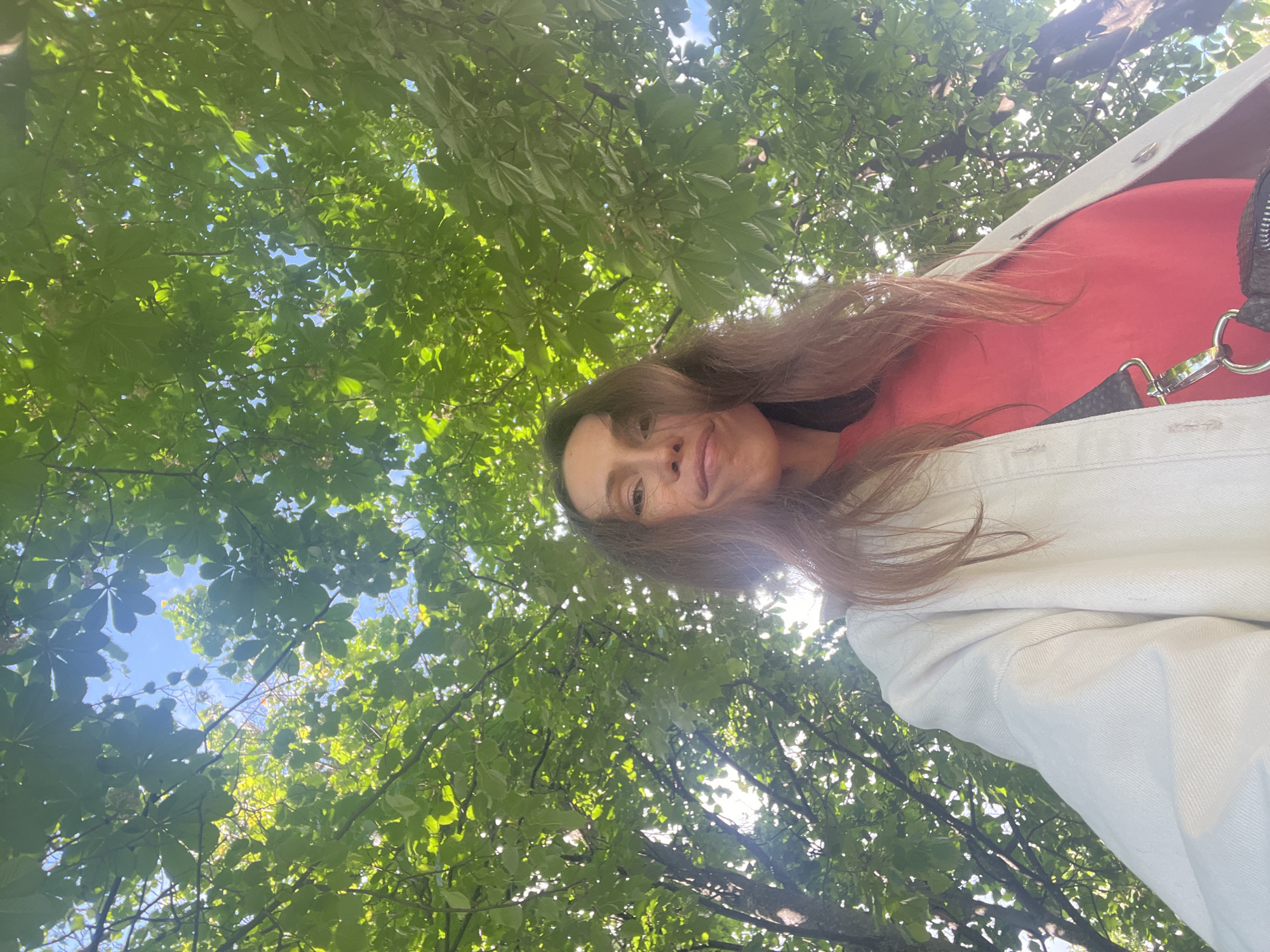 The image is a unique low-angle portrait, rotated 90 degrees counter-clockwise, featuring a woman looking down towards the camera from an upward perspective. She has long, flowing dark brown hair and visible freckles on her face, with a gentle, slightly smiling expression. She is dressed in a red top, layered with a light-colored (likely white or off-white) jacket or coat. A black bag strap with a prominent silver clasp is visible near her shoulder.
The background is filled with a dense canopy of lush green leaves from deciduous trees, through which patches of clear blue sky are visible. Sunlight filters through the foliage, creating bright spots. This indicates an outdoor setting, likely a park or garden in Moscow, Russia, during daytime under clear or partly cloudy weather. There is no visible text in the image.