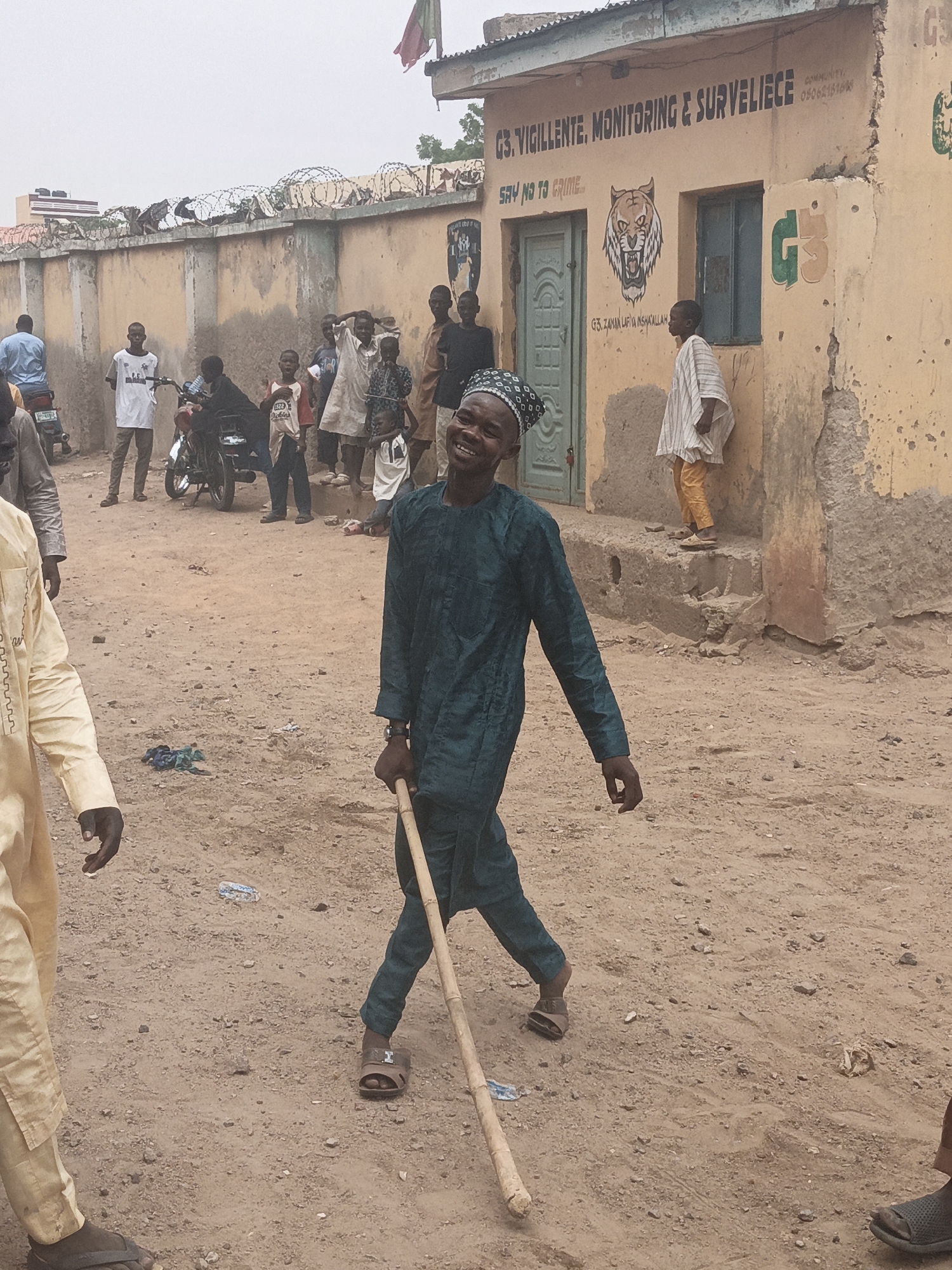 The image depicts a street scene in Gwagwarwa, Nigeria. In the foreground, a man wearing a dark teal tunic and pants, and a patterned skullcap, is walking towards the right, holding a wooden staff. He is smiling broadly. To his left, another man in a light yellow tunic and pants is partially visible. In the background, several other individuals, including children and adults, are gathered. A motorcycle is parked on the left side of the frame. Behind the people, a building with a pale yellow stucco exterior displays signage that reads "G3. VIGILENTE, MONITORING & SURVELIECE" and "SAY NO TO CRIME". The ground is sandy and uneven, with scattered debris. The sky is overcast.