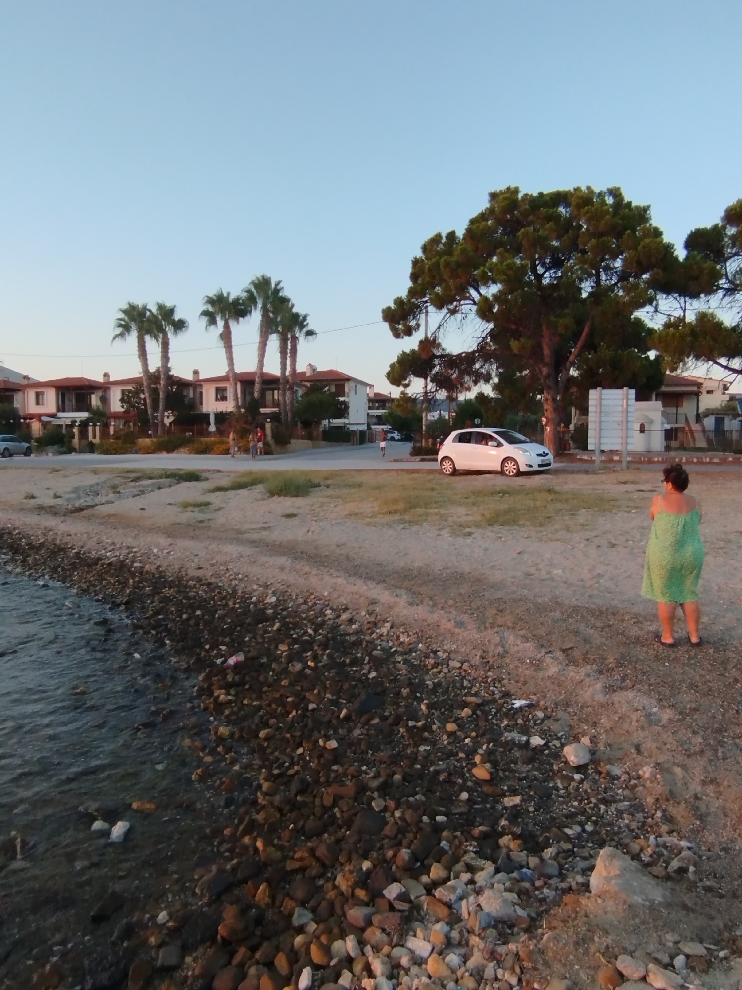 A medium shot captures a beach scene in Sithonia Municipal Unit, Greece, at dusk. The foreground features a rocky shoreline meeting the calm, dark water of the sea. The beach transitions to sand further inland, with scattered debris visible.
In the mid-ground, a woman stands facing away from the camera, wearing a green dress and flip-flops. She is on the sandy part of the beach, near a paved road. A white car is parked on the road to her left, with a large, mature pine tree dominating the background to her right.
Behind the road, a row of houses with red-tiled roofs and white facades is visible, with palm trees interspersed. The sky is clear and a light blue, indicating the approaching evening. Two figures are walking on the road in the distance, and another car is parked further down the road. The overall impression is a quiet, seaside location at the end of the day.