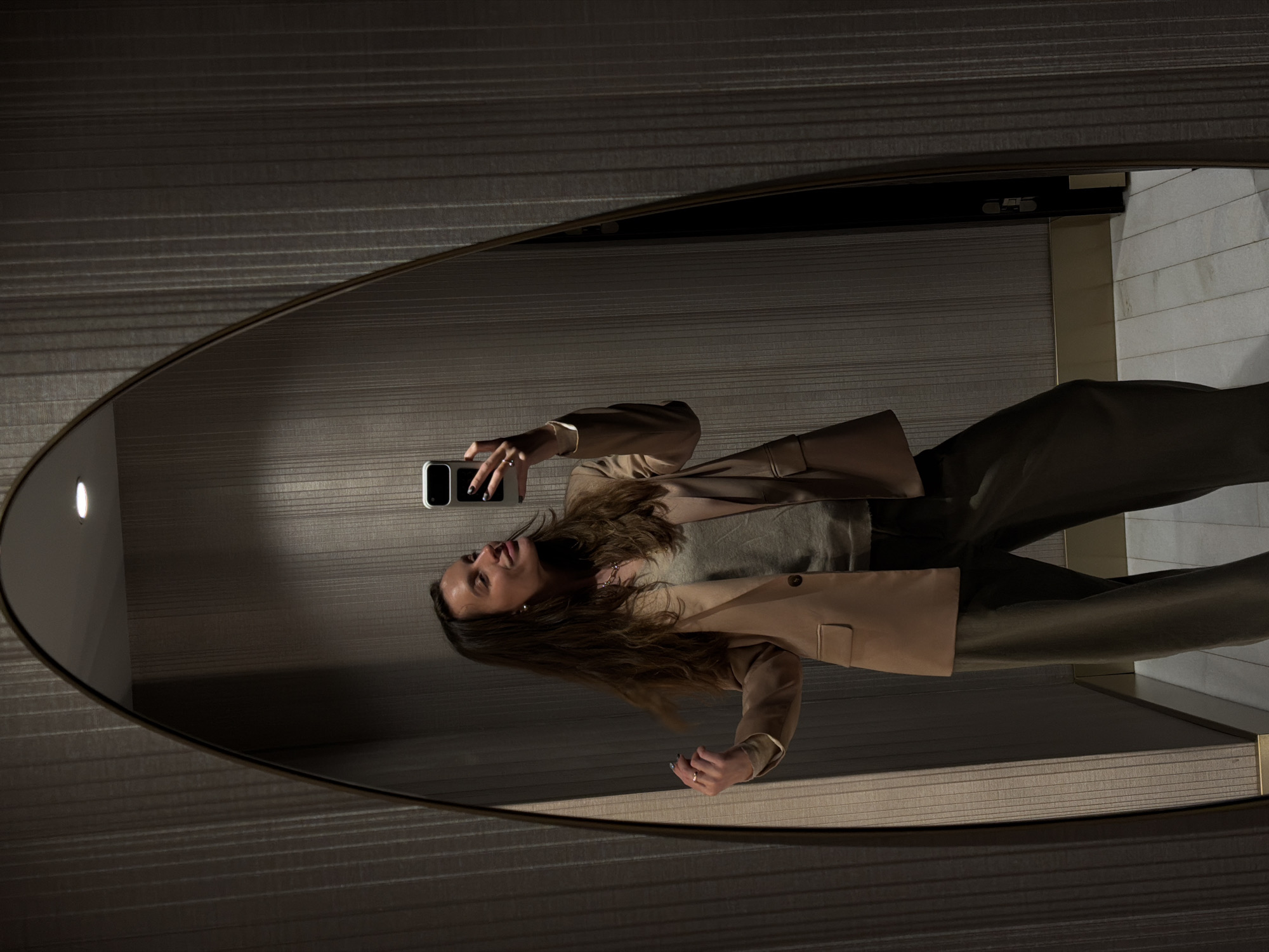 A person is reflected in an oval mirror. The person is holding a cell phone and looking towards the camera. They are wearing a blazer, a top, and pants. The background consists of patterned wallpaper and a portion of a wall, adjacent to a door, which appears to be leading to a bathroom. The scene is indoors, and based on the design style, it might be a hotel bathroom. The location in Budva, Montenegro, cannot be verified from this image.