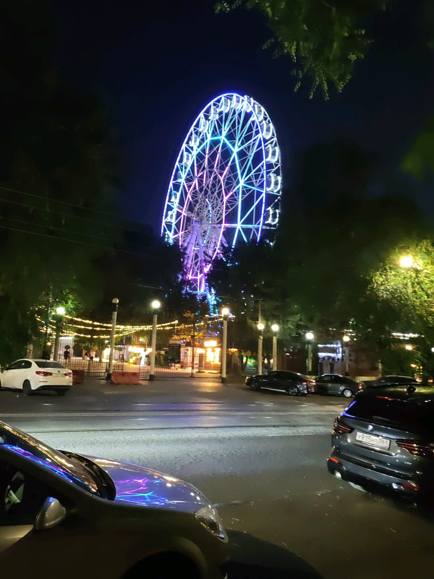 This image depicts a nighttime scene in Rostov-on-Don, Russia, likely in an amusement park or public space illuminated by a Ferris wheel and festive lights. The large Ferris wheel is prominently displayed, adorned with a vibrant mix of blue and pink lights, suggesting it is operational. In the foreground, several cars are parked or driving on a street. One car, a white sedan, is visible on the left side of the road. To the right, a dark-colored SUV with a Russian license plate is parked. The area appears to be a pedestrian zone or entrance to an attraction, with a fence and scattered streetlights and string lights adding to the ambiance. Trees frame the scene, their foliage dark against the night sky. The ground appears to be paved, with reflections of the lights visible on the wet surface of the road and car hoods. People are visible in the background, suggesting the presence of visitors or staff in the illuminated area behind the fence.