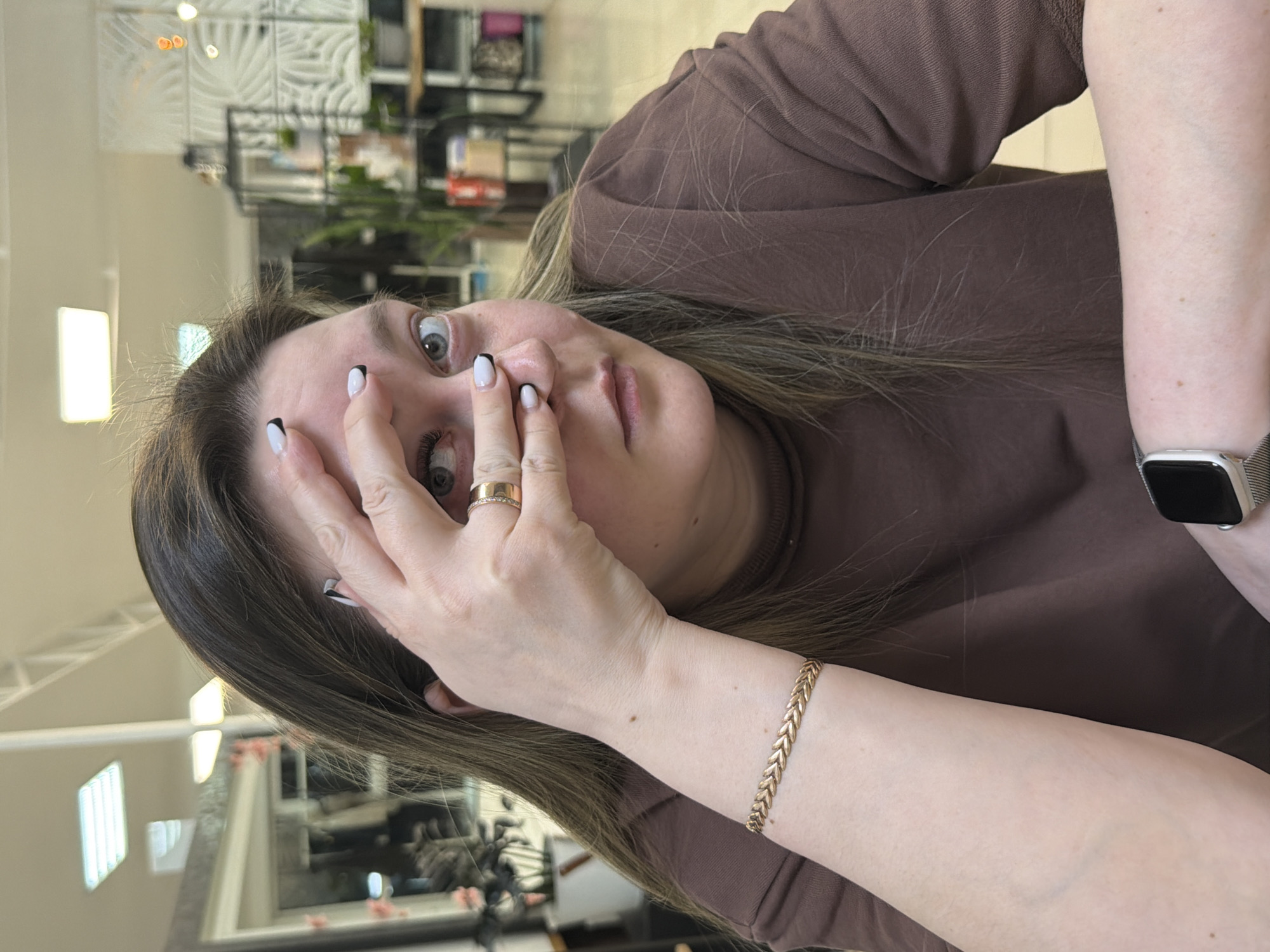 A woman is depicted from the chest up, positioned diagonally across the frame with her head tilted. She has light brown hair, which is styled down, and wide-open grey eyes. Her expression conveys surprise or mild astonishment. Her left hand is raised to her face, partially covering her mouth and lower nose, with her fingers slightly spread. She sports white nails with black tips, resembling a French manicure style, and a gold ring with small embedded stones on her left ring finger. A gold-colored bracelet with a chevron or herringbone pattern adorns her left wrist, and a silver-colored smartwatch with a light band is visible on her right wrist. She is wearing a simple brown long-sleeved top or sweatshirt.
The background shows an indoor commercial setting, possibly a salon or shop, with soft lighting. Details include blurry shelves, possibly displaying products, and decorative elements like geometric wall panels and reflections in mirrors. The overall scene appears candid, capturing a spontaneous moment. There is no visible text in the image.