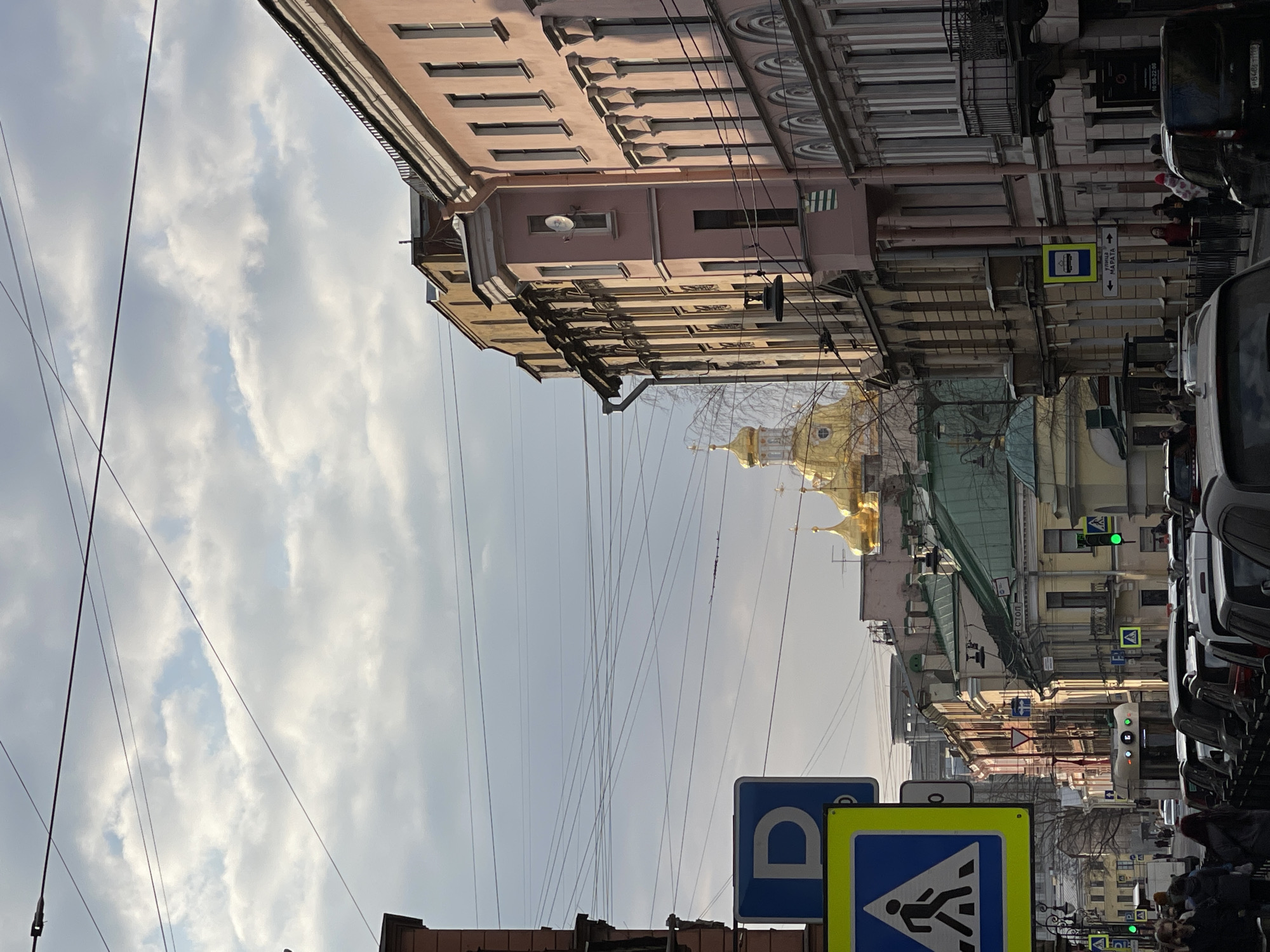 The image captures a street scene in Saint Petersburg, Russia, viewed from a low angle looking down the street. The architecture is characterized by ornate buildings, with a pink building visible on the left side of the image. The sky, which dominates the upper portion, is filled with clouds. Power lines crisscross the space above the street. The golden dome of the St. Nicholas Naval Cathedral is partially visible in the distance. Street signs are present in the foreground, and parked cars line the right side of the street. Pedestrians and other vehicles are visible further down the street.