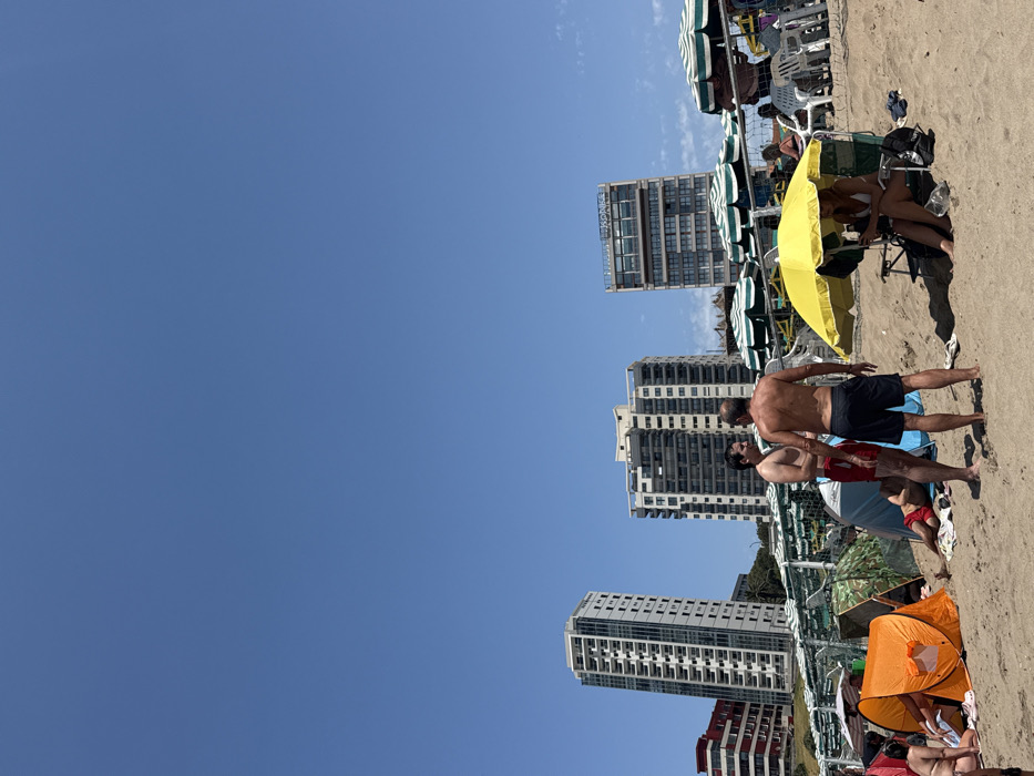 A sunny beach scene with several high-rise buildings in the background. People relax under beach umbrellas and in small pop-up tents. A group of shirtless men stand on the sand in the foreground. The clear, blue sky takes up a large portion of the image.