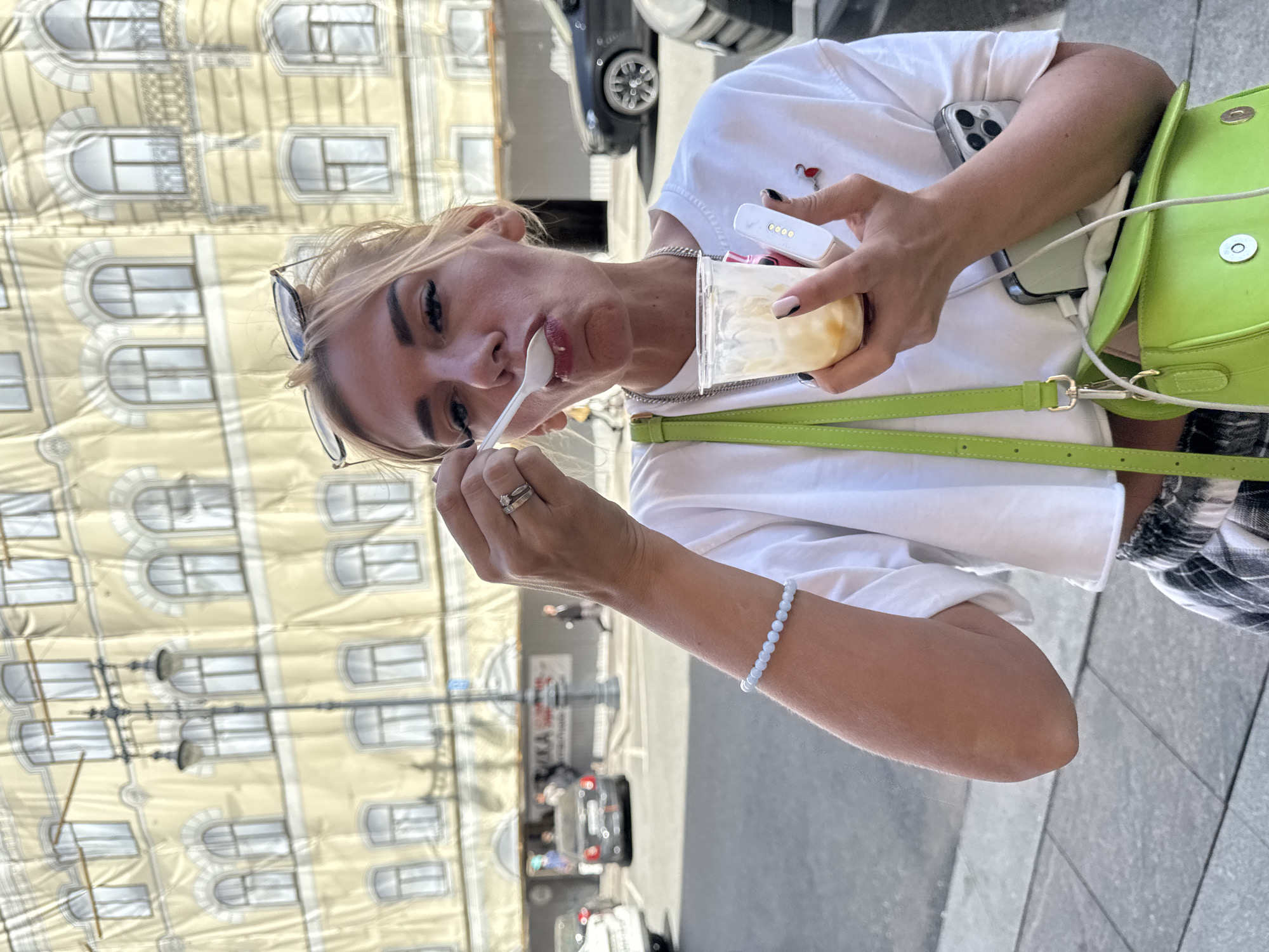 A young woman with blonde hair is captured in a close-up shot, looking at the camera while eating from a clear plastic cup with a white spoon. She is wearing a white t-shirt and sunglasses pushed up on her head. A lime green purse with a strap is visible across her body, and she is holding a white electronic device in her right hand. Her left hand, adorned with a ring, holds the spoon to her mouth.
The background is slightly out of focus, showing a light yellow building with numerous windows. The image appears to be taken during the daytime, under sunny weather, given the bright lighting and shadows. The presence of cars in the background and the architectural style of the building suggest an urban setting, specifically Saint Petersburg, Russia. The overall mood is casual and relaxed, with the woman seemingly enjoying her treat while on the go.
The woman has a light blue beaded bracelet on her left wrist. On her white t-shirt, there is a small red embroidered flamingo pin. A silver chain necklace is also visible around her neck. A smartphone is tucked into her shirt. The image is rotated 90 degrees counterclockwise.