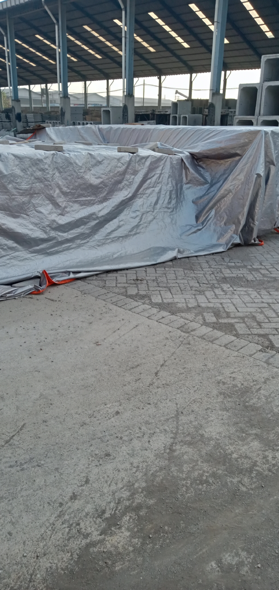 The image depicts a large silver tarp covering a stack of rectangular concrete blocks in an industrial setting. The scene is under the roof of a large open-sided building with steel support columns and a metal roof with rectangular skylights. In the background, more concrete blocks are visible. The foreground shows a concrete surface transitioning to a brick-paved area near the tarp. The tarp is secured with small concrete blocks. The location appears to be an outdoor storage area for precast concrete materials, possibly a manufacturing or construction supply yard. No people or other subjects are visible in the image.
