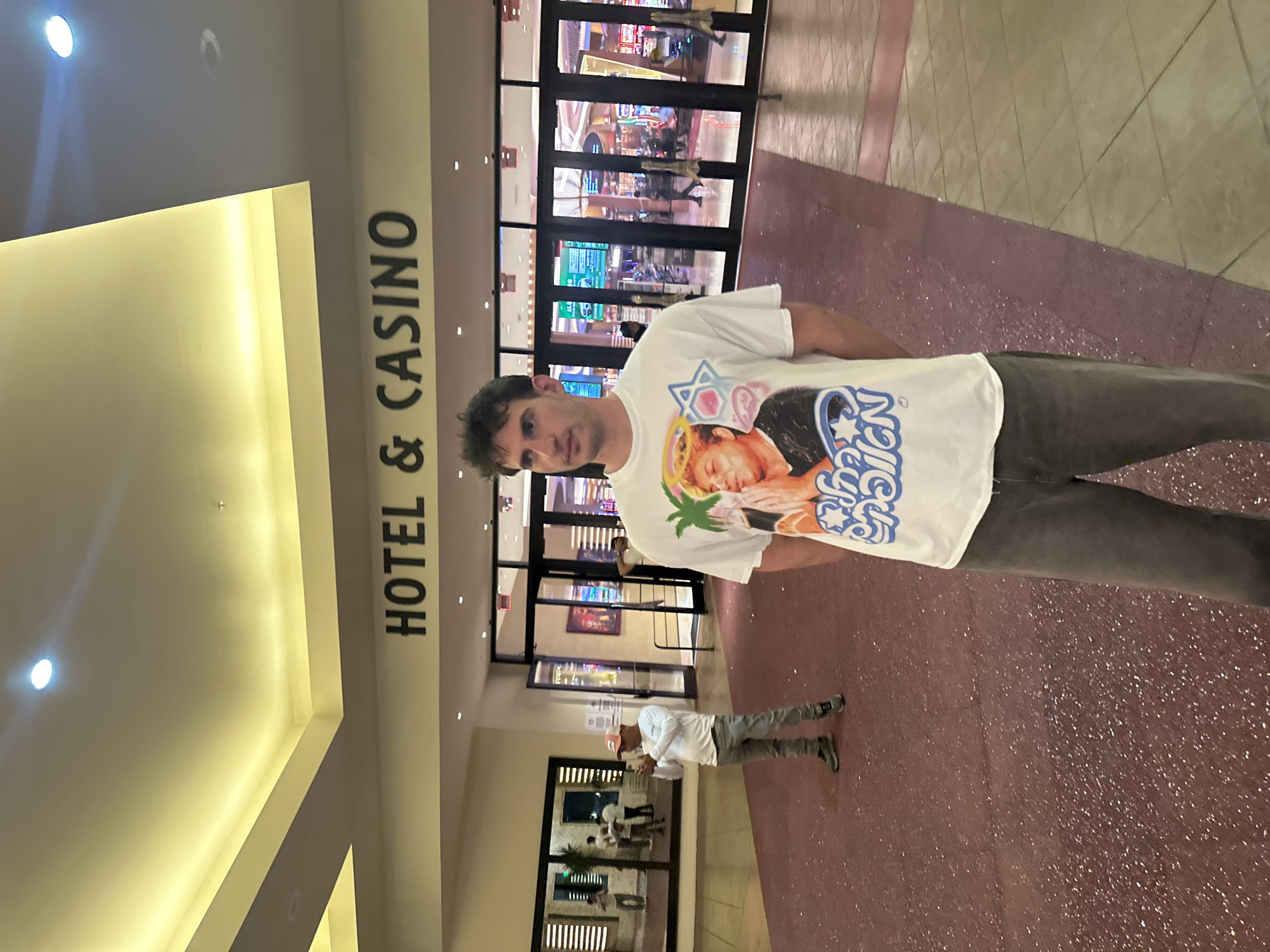 The image depicts a man standing in what appears to be the entrance of a hotel and casino. The words "HOTEL & CASINO" are prominently displayed on a column to his left. The interior of the casino is visible through glass doors behind him, showcasing various gaming machines and people. The man is wearing a white T-shirt with a graphic design and grey pants. Another person is seen walking behind the man. The scene is located in Hollywood Seminole Reservation, United States of America.