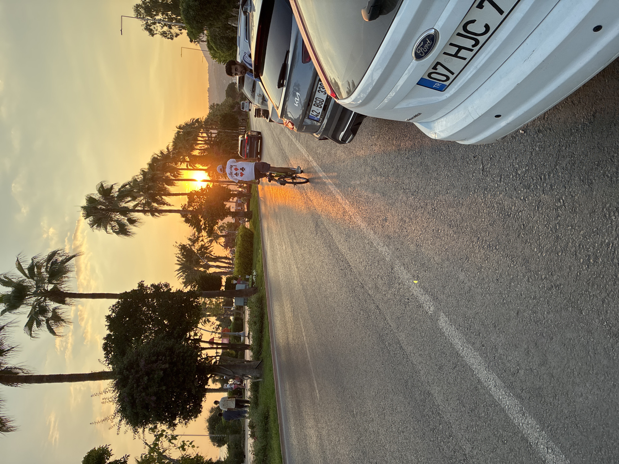 The image captures a street scene in Alanya, Turkey, during sunset, with the sun low on the horizon casting a warm glow. A person is cycling on the right side of the road, facing away from the camera. Several cars are parked along the right side of the road, including a white Ford with a license plate that reads "TR 07 HJC 7". To the left of the road, there is a sidewalk lined with palm trees and other foliage, with several people walking. The sky is orange and yellow, indicative of a sunset. The road surface appears to be asphalt with a white lane marking.
