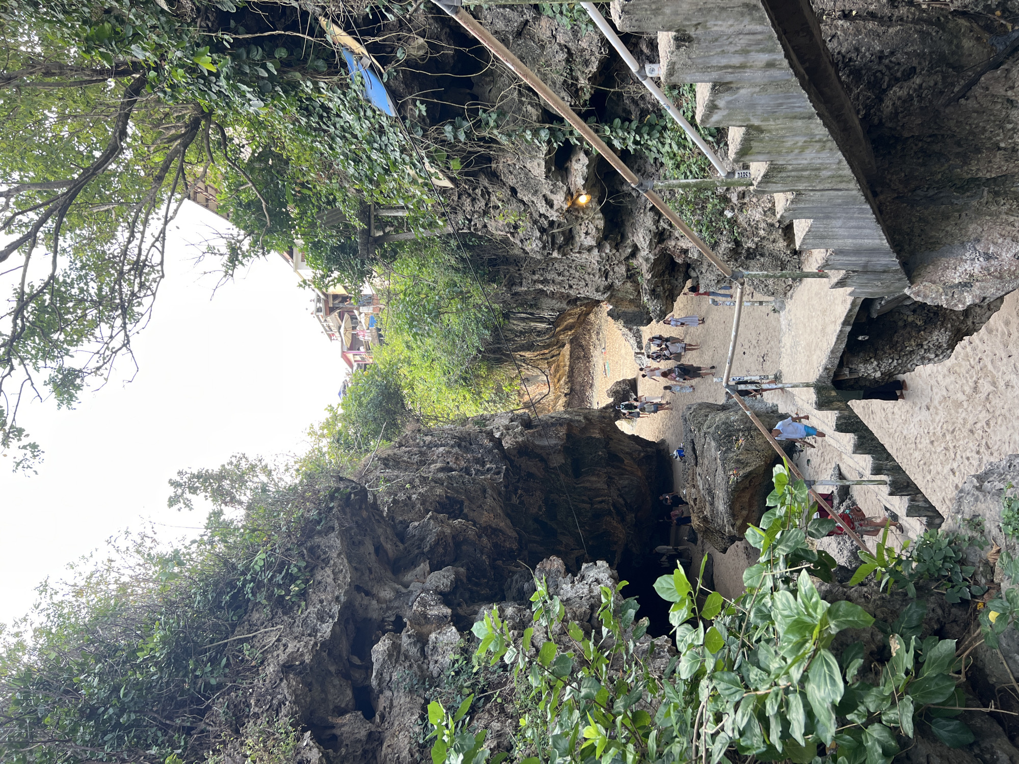 In Kuta Selatan, Indonesia, this image captures a scene at a beach with a dramatic geological formation. A large rock face, weathered and covered in lush green vegetation, dominates the left side of the frame. On the right, a set of concrete stairs with sturdy railings descends towards a sandy area. The stairs are worn, showing signs of age and use.
The beach itself is characterized by fine sand and is partially enclosed by the imposing rock structures, creating a cave-like entrance in the distance. Several people are visible on the beach and on the path leading into the rocky enclosure. They appear to be tourists, casually dressed and walking around, some in groups, exploring the area. A few individuals are seated on the sand.
Beyond the immediate beach area, hints of human habitation can be seen, with buildings perched on the cliffs above, some with umbrellas suggesting a tourist establishment. The overall environment suggests a popular tourist destination where natural beauty, including striking rock formations and a beach, is combined with accessibility provided by man-made structures like stairs. The lighting indicates it is daytime, with overcast skies creating a soft, diffused light.