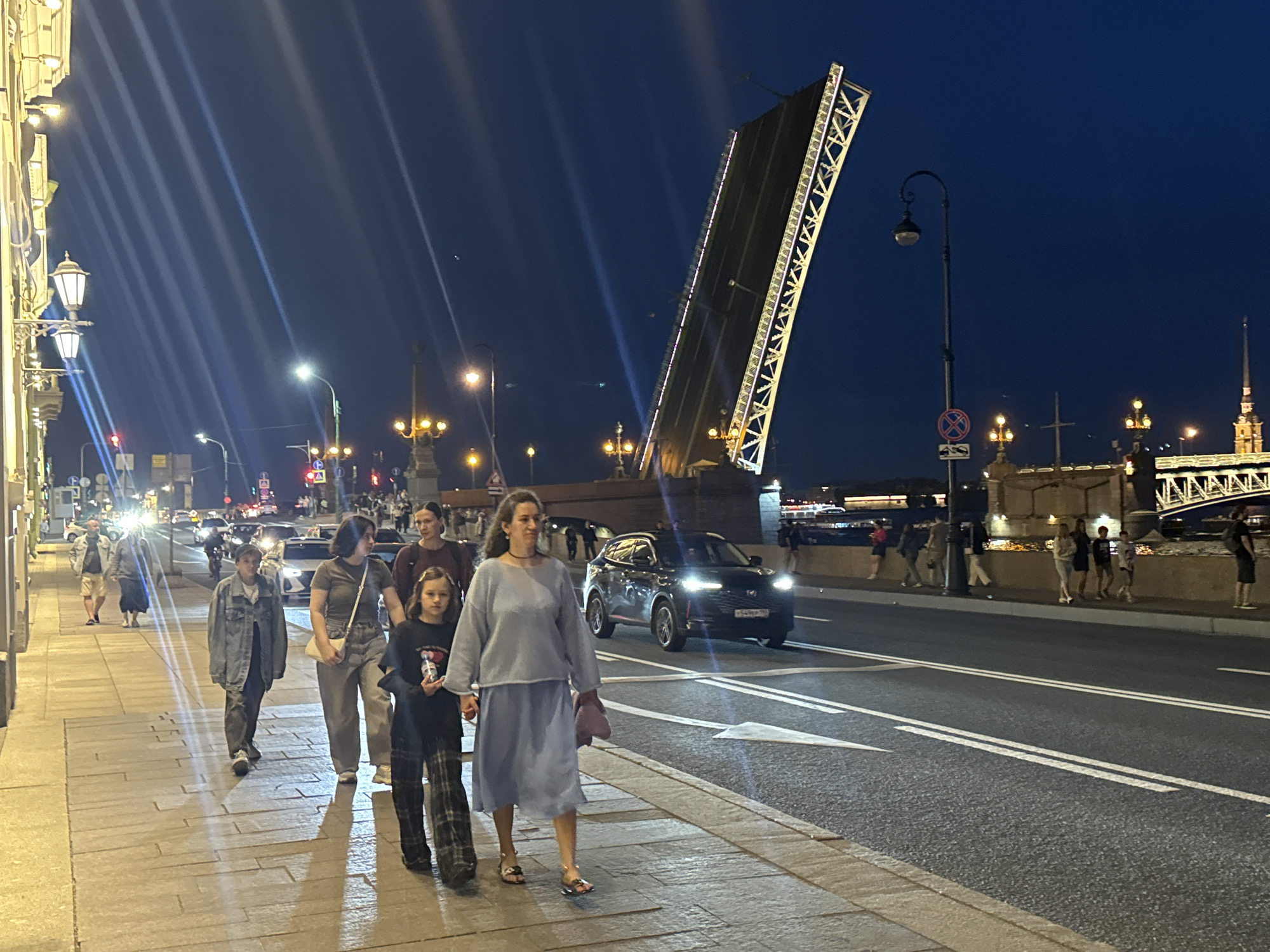 This nighttime image captures a scene on the Admiralty or Palace Embankment in Saint Petersburg, Russia, looking east-northeast towards the raised Palace Bridge (Dvortsovy Most) over the Neva River and the illuminated spire of the Peter and Paul Cathedral in the distant background.
In the immediate foreground, a paved sidewalk on the left is illuminated by warm yellow light from ornate streetlights mounted on a classical-style building facade. Seven pedestrians are visible on this sidewalk, casting long shadows to their right. From left to right, individuals include one in a light jacket and shorts, another in a dark skirt and light top, and a person in a denim jacket and dark pants. Closer to the viewer, a woman in a light blue sweater and skirt holds the hand of a child in a dark shirt and plaid pants, with another woman in a maroon top positioned behind them. In the mid-ground, a two-lane asphalt road features a solid double white line and a painted white directional arrow. A dark-colored Chery Tiggo 8 Pro SUV, with its headlights on and a Russian license plate (e.g., P549PE), drives away from the viewer in the right lane.
The dominant feature in the mid-ground and right is the Palace Bridge, with both of its central drawbridge sections fully raised at a steep angle. The undersides of these sections display illuminated white structural trusses along their edges. To the right of the road, a pole bears a red and white "no stopping" sign. The background reveals a deep blue night sky. Distant city lights and structures are visible across the river on the far embankment, where several more people are discernible. The illuminated spire of the Peter and Paul Cathedral stands prominently against the sky in the far distance on the right.