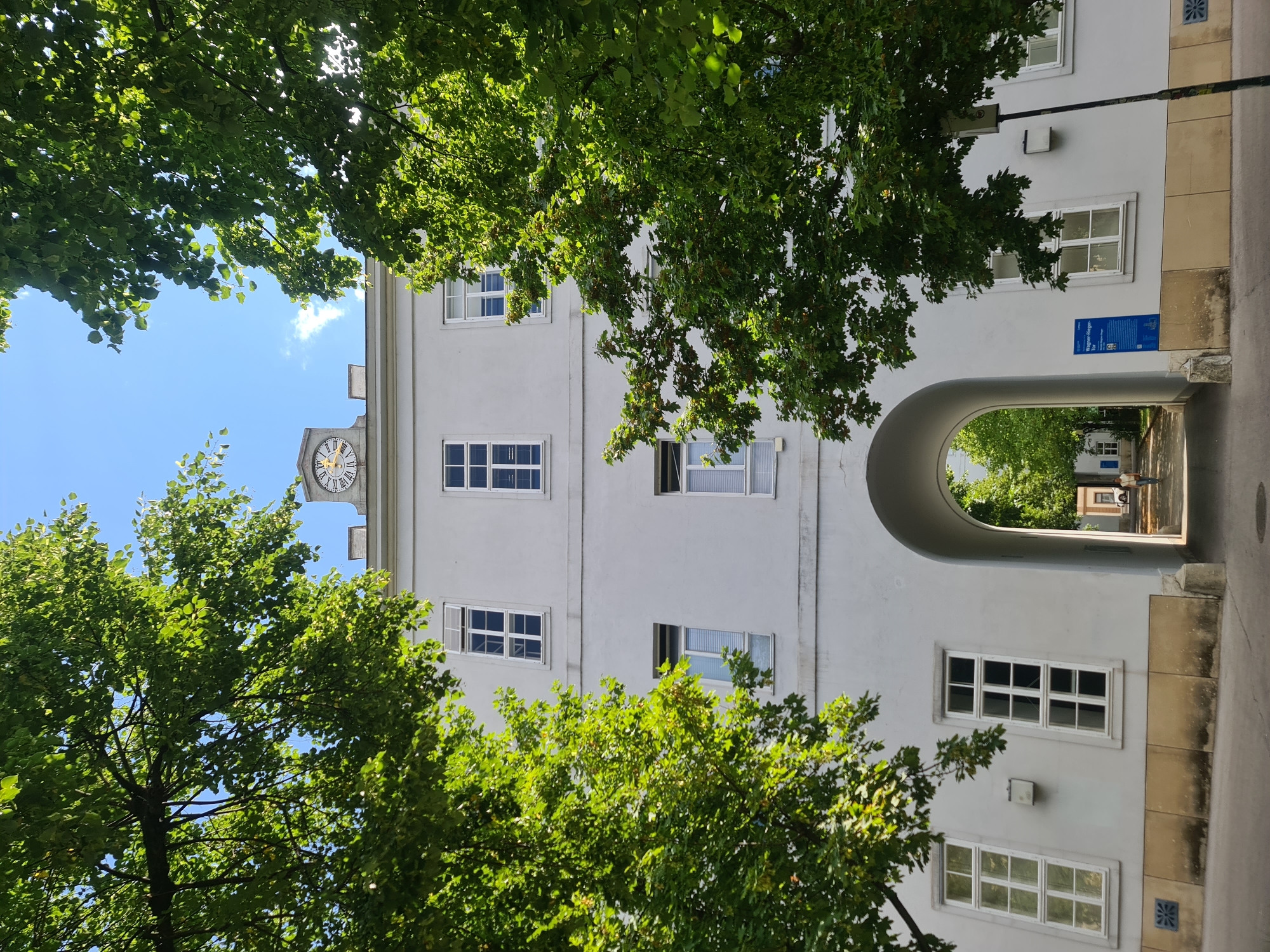 The image shows the Wagner-Jauregg-Tor in Vienna, Austria. The architectural structure features a large, arched entranceway leading into a courtyard. The building is a light grey color with multiple white-framed windows on its facade. A clock with Roman numerals is visible at the top of the building, indicating the time. Lush green trees frame the scene, with their branches extending into the bright blue sky. A person is walking through the arched entranceway into the courtyard, where more trees and buildings can be seen in the distance. A blue informational plaque is mounted on the wall near the archway. The ground in front of the building appears to be paved.