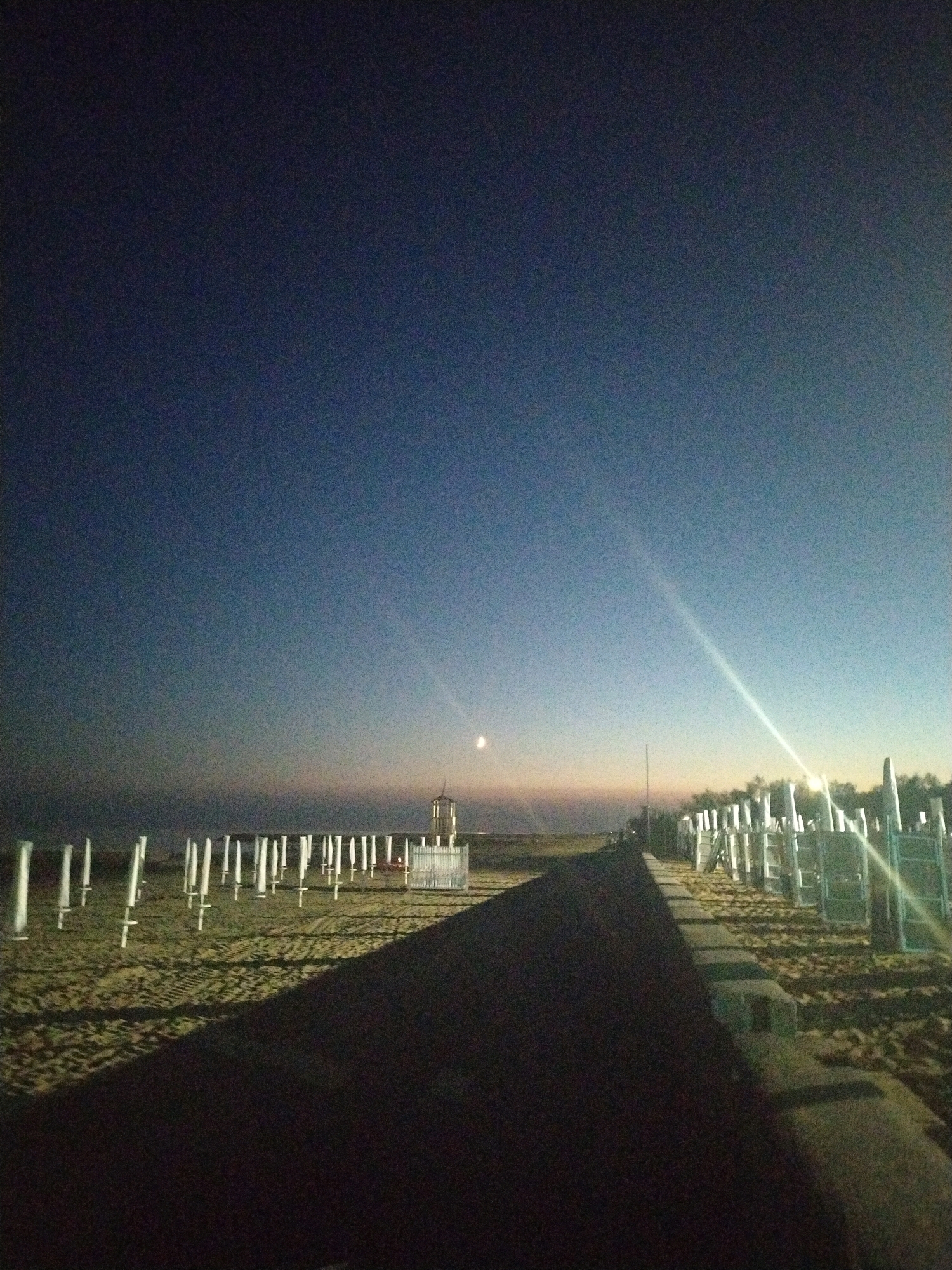 The image depicts a beach scene at twilight in Caorle, Italy. A paved walkway runs diagonally across the foreground, casting a long shadow to the left. Beyond the walkway, rows of tall, white posts are arranged on the sandy beach. In the distance, a small, open-air structure resembling a lifeguard tower or pavilion is visible. The sky is a deep gradient of blues and purples, with a faint moon visible near the horizon. The overall atmosphere is serene and quiet, suggesting an evening setting. The presence of the beach, walkway, and posts indicates a public or recreational area. There are no people visible in the image.