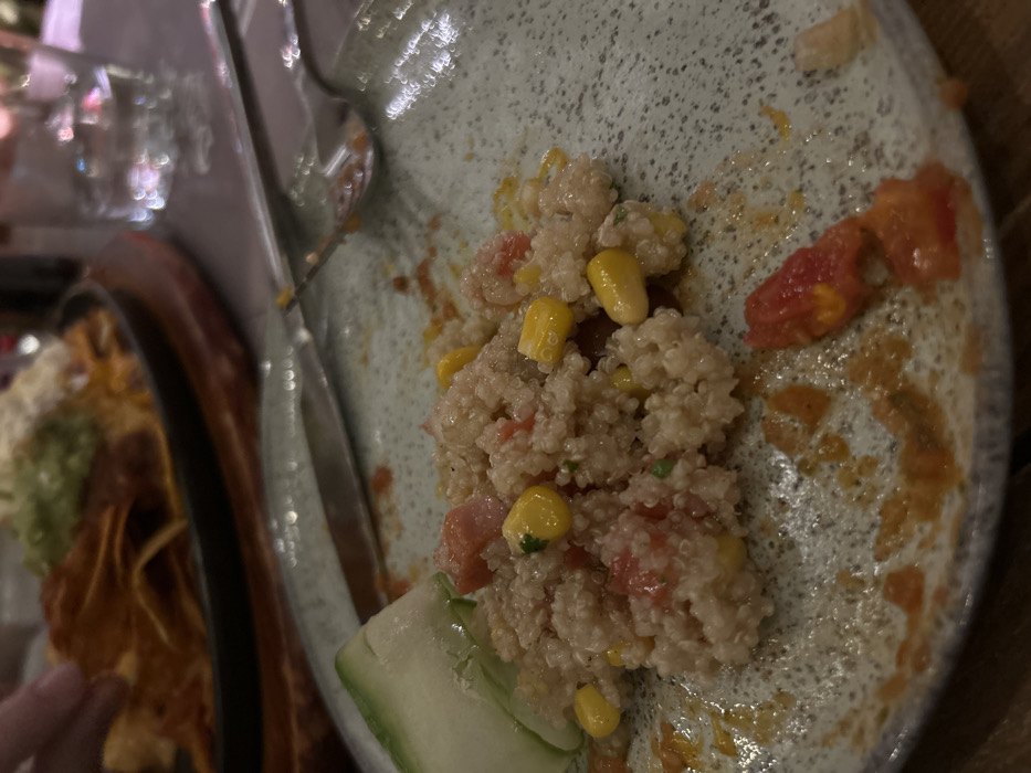 This close-up image shows a partially eaten meal served on a speckled, light-colored plate. The main food item visible is a quinoa salad, mixed with bright yellow corn kernels and diced red tomatoes. A slice of green cucumber rests on the side of the plate. Reddish sauce or dressing residue is spread across the plate, indicating the food has been consumed.
To the left, out of focus, another dish holds a pile of golden-brown tortilla chips (nachos) accompanied by a dollop of green guacamole or avocado dip. A person's hand is visible in the lower-left corner, reaching towards the nachos. A metal fork lies on the main plate.
The lighting is dim and artificial, creating reflections of blurred objects like glasses or bottles in the background, suggesting an indoor dining setting, likely in the evening or at night. The scene takes place in Melissia, Greece. No text is visible in the image.