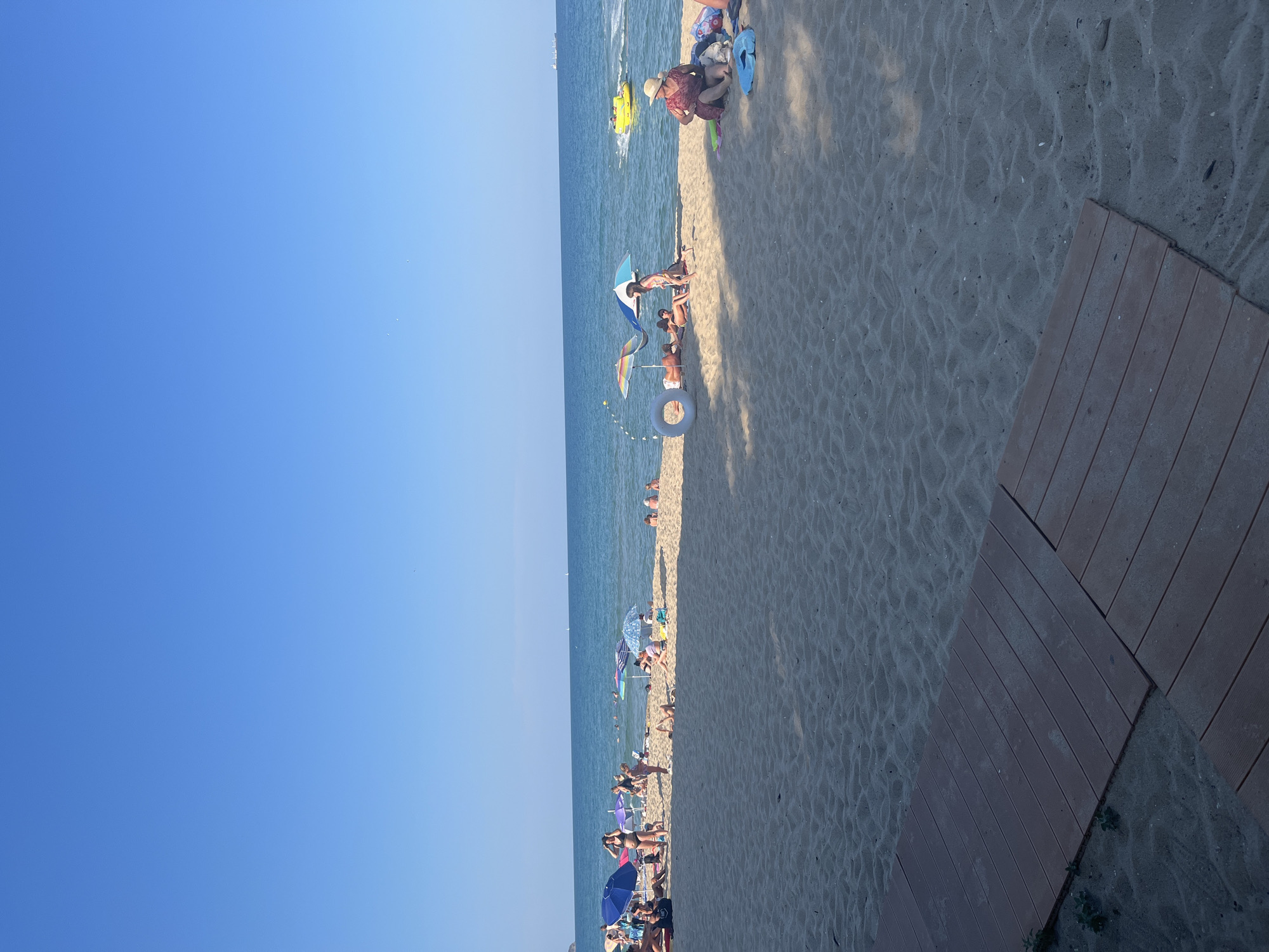 The image depicts a sunny day at a crowded beach in Obzor, Bulgaria. In the foreground, a wooden boardwalk is laid on the sand, leading towards the sea. The beach is populated with numerous people relaxing under umbrellas, sunbathing, and swimming in the clear blue water. Several beach umbrellas of various colors, including blue, purple, and a rainbow-striped one, are scattered across the sand. Children and adults are visible in the water and on the shore. A yellow inflatable tube and a white inflatable ring are present near the water's edge. In the distance, a yellow inflatable watercraft is seen on the sea, and a large ship is visible on the horizon. The sky is a clear, cloudless blue. The overall scene conveys a typical summer beach atmosphere.
