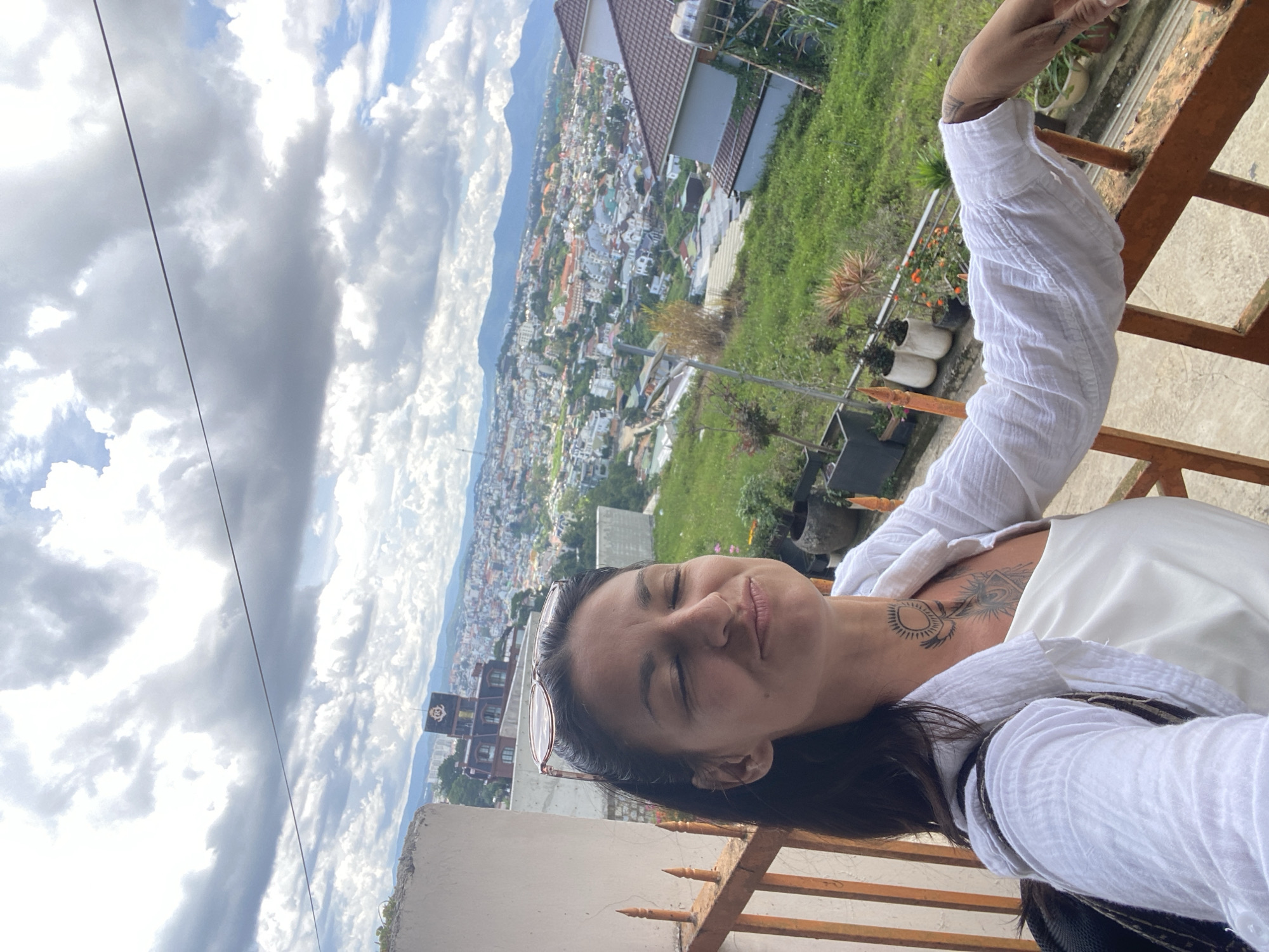 A woman with dark hair and a tattoo on her neck is taking a selfie on a balcony overlooking the city of Da Lat, Vietnam. The woman is wearing a white shirt over a white top. Her eyes are closed, and her mouth is pursed. She is leaning on a rusty orange railing. In the background, the city of Da Lat sprawls across rolling hills. Buildings of various colors and sizes are visible, nestled amongst lush green vegetation. Mountains form a backdrop to the city, under a sky filled with dramatic clouds. Several potted plants are arranged on the balcony and the slope below. A building with a dark clock tower and a distinctive architectural style is prominent on a hill in the mid-ground.