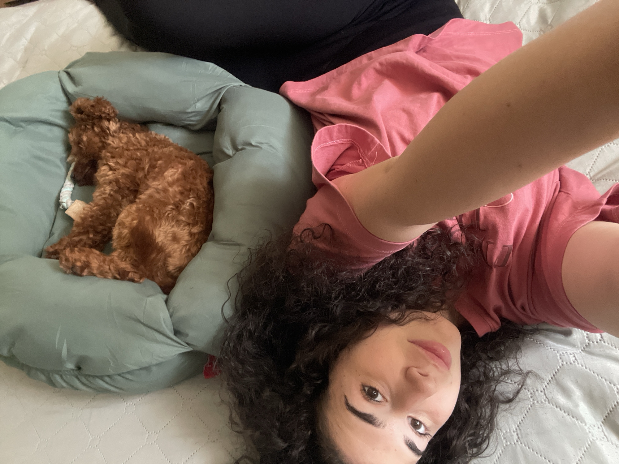 A young woman with dark curly hair and a pink t-shirt is lying on a gray quilted surface next to a green dog bed with a brown poodle in it. The woman's right arm is raised in the air. Behind her head is another dark-colored fabric, possibly pants. The location is likely indoors and difficult to determine as Sofia, Bulgaria, without further context.