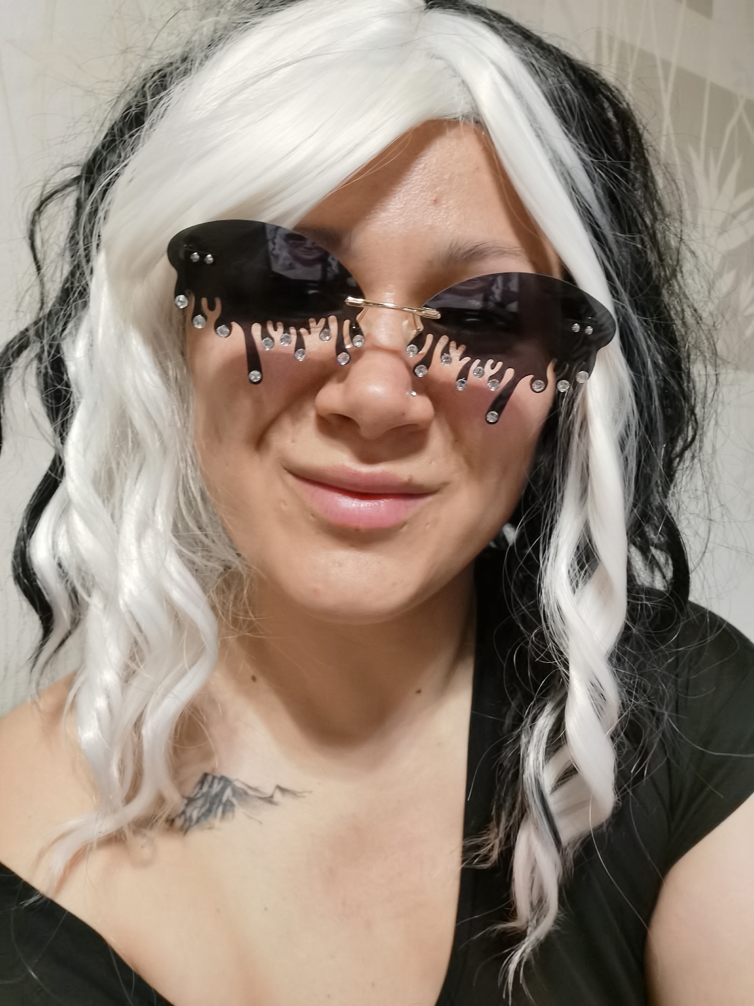 A person with fair skin is centered in the frame, wearing sunglasses with a black drip design and small, clear gems. They have a black and white wig with wavy strands. A tattoo of a mountain range is visible on their upper left chest. They are wearing a black garment. The background is a blurred, neutral tone. The location and city are not discernible from the image.