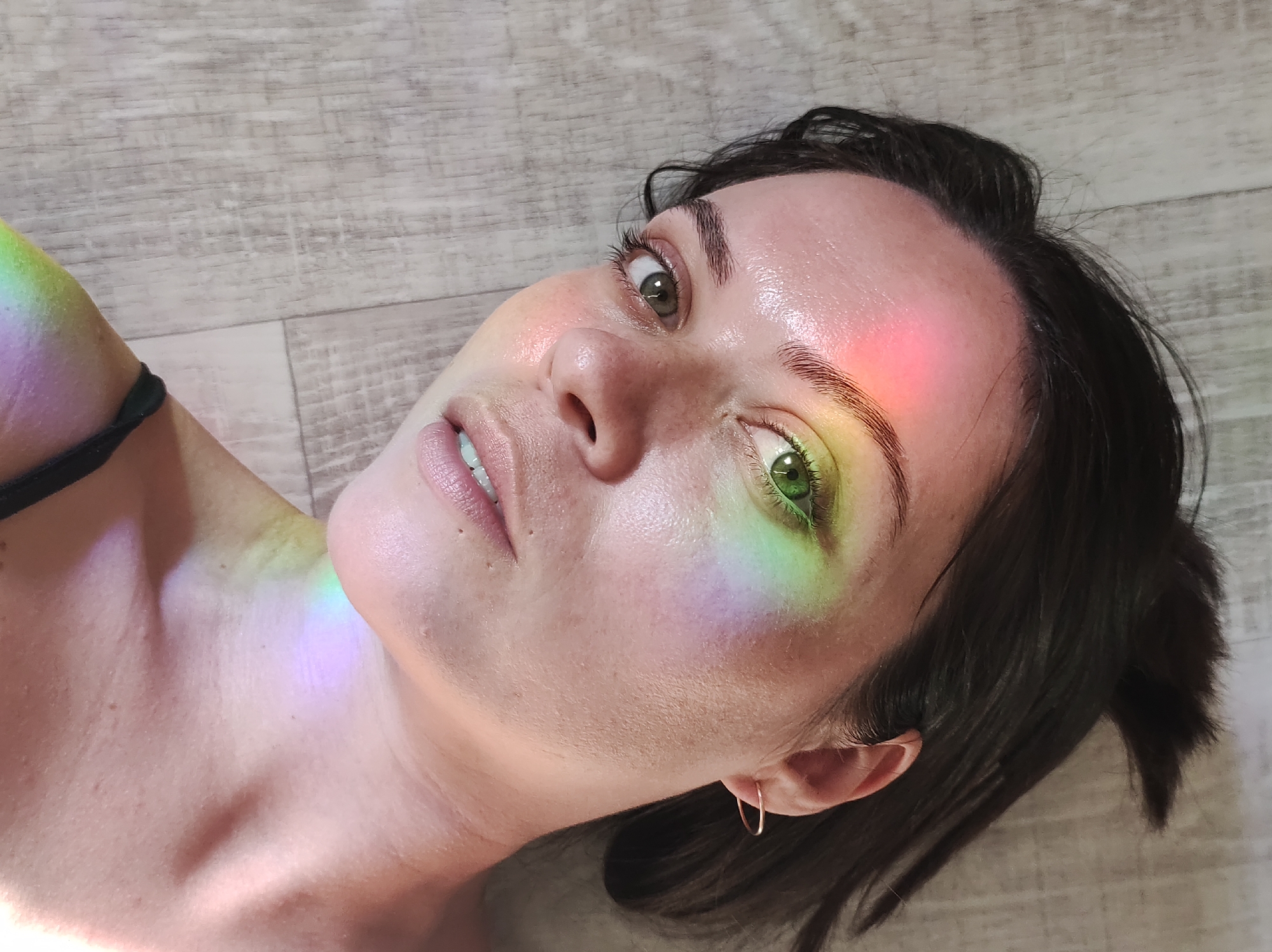 A close-up, slightly angled shot depicts a fair-skinned individual with dark hair, light green eyes, and a gold hoop earring in their left ear. The individual is lying down on what appears to be a wood-patterned surface. A prism of rainbow light is cast across their face and upper chest, illuminating their forehead, nose, left eye, cheek, and shoulder with varying colors. The location in the city of Unknown, Unknown is not identifiable from the given information.