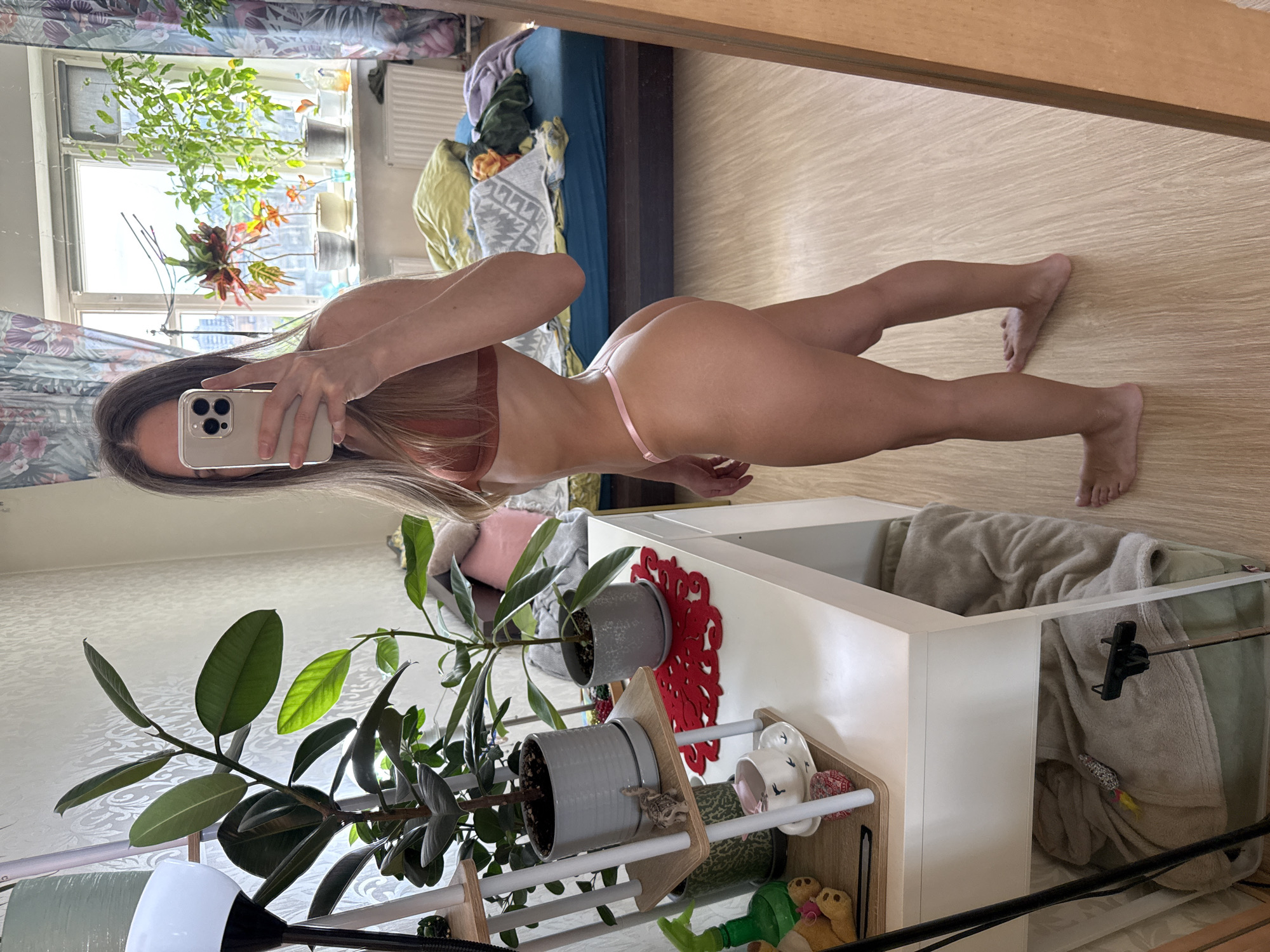 A woman is taking a mirror selfie in a room. She is standing next to a white desk with plants on it. The room appears to be a bedroom, with a bed in the background. The woman is wearing a bikini and is looking at her phone. The lighting is natural, coming from a window. The image is taken from a slightly low angle.