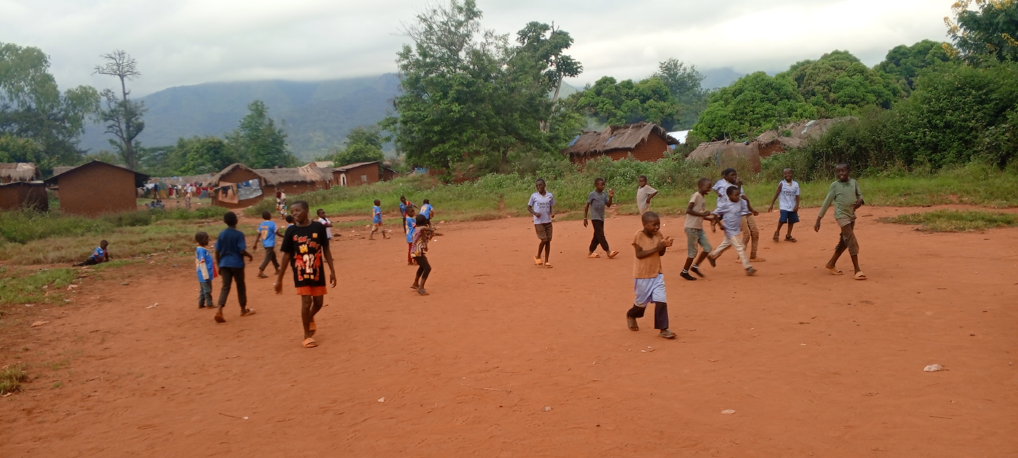 In a rural area of Ngalula, Democratic Republic of the Congo, several children are engaged in play on a reddish-brown dirt surface. The background reveals a village setting with modest, possibly mud-brick homes that have thatched roofs. The sky appears overcast, and in the distance, there are forested hills or mountains. Some children are walking, while others seem to be engaged in a game or other activity. The attire of the children varies, with some wearing t-shirts and shorts and others in more casual clothing. A few of the children are smiling.