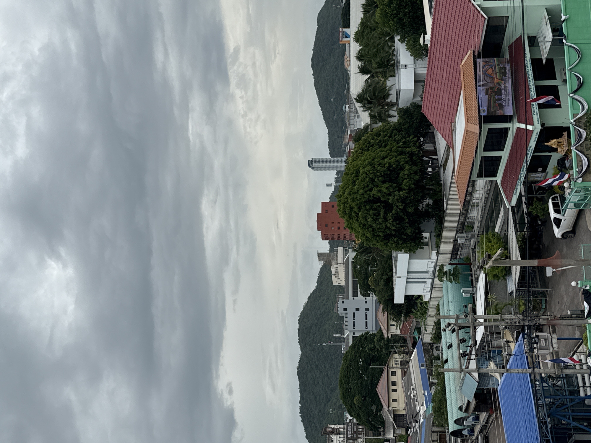 This is an aerial view of a city, likely in Hua Hin City Municipality, Thailand, under a cloudy sky. In the foreground, there are several buildings with various colored roofs, including red, blue, and green. Utility poles and electrical wires are visible, along with some trees. A white pickup truck is parked on the right side of the image. In the mid-ground, more buildings and trees are present, with a large green tree prominently displayed. The background features lush green hills and mountains, with some buildings and a tall skyscraper visible. The overall scene depicts an urban landscape interspersed with natural elements.