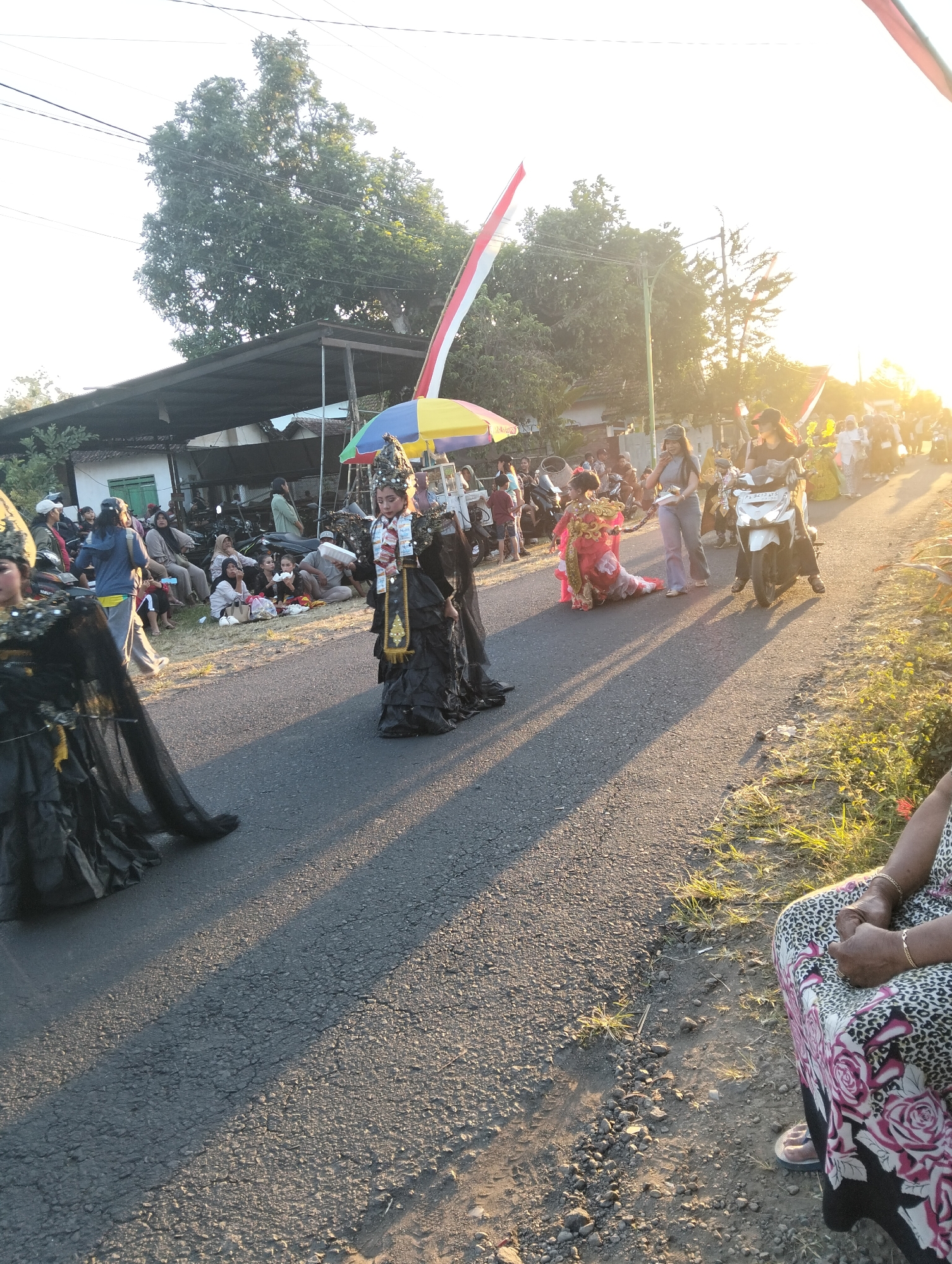 A street parade is in progress in Sumbersewu, Indonesia, as indicated by the Indonesian flag prominently displayed. The event appears to be taking place in the late afternoon or early evening, with the low angle of the sun casting long shadows across the asphalt road. The foreground features participants in ornate costumes, including a person in a black, ruffled outfit and elaborate headwear, and another in a red and gold traditional garment. Several motorcycles are present, some parked along the side of the road and others being ridden as part of the procession. Spectators are gathered on the side of the road, some sitting on the ground and others standing. The environment includes trees and buildings lining the street, suggesting a semi-rural or town setting. The overall atmosphere seems festive, likely related to a cultural celebration or event.