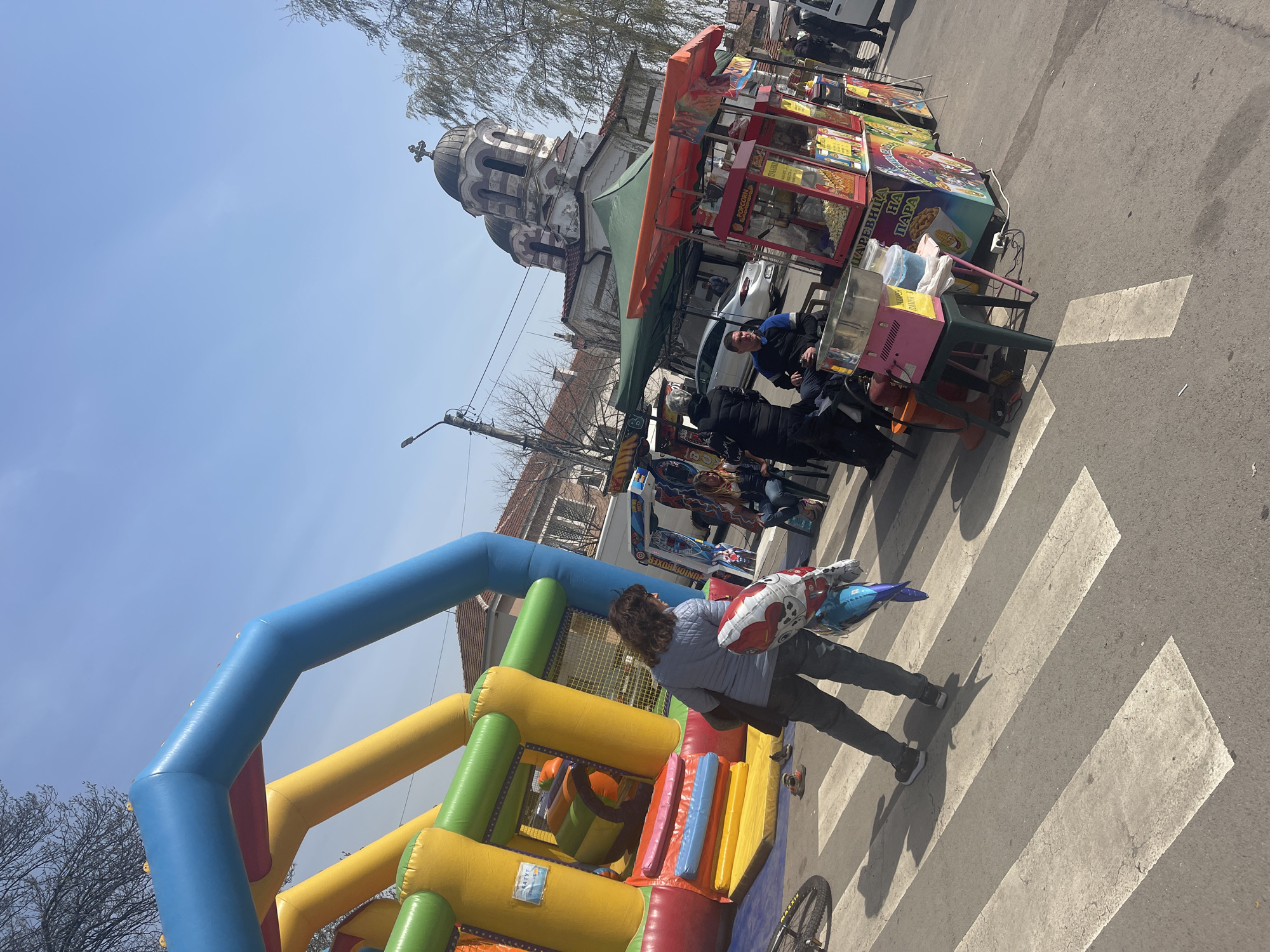 The image shows an outdoor scene on a street in Sofia, Bulgaria. In the foreground, there is an inflatable bouncy castle with blue, yellow, green, and red sections. A person is walking across a zebra crossing, carrying a balloon. In the background, there are various food stalls and vending carts. A church with domed towers is visible behind the stalls. Several people are standing around the vending carts. The sky is blue and the weather appears to be sunny.