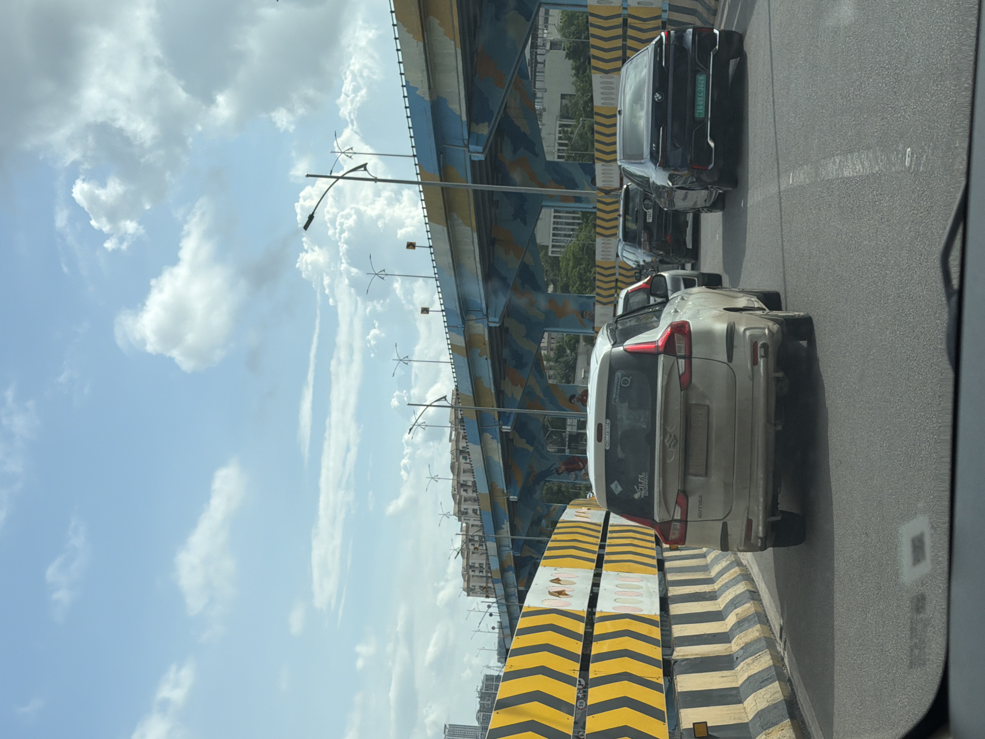 The image shows a traffic scene on an elevated highway in Hyderabad, India. In the foreground, a beige colored MPV is in the left lane, partially obscuring a dark-colored sedan in the right lane. The sedan has a license plate that reads "TG09L302M". In the middle ground, several other vehicles are visible, including a black SUV. The highway is bordered by a yellow and black striped barrier. In the background, a multi-colored elevated bridge with abstract patterns in blue and yellow spans across the scene. The sky is partly cloudy with scattered white clouds against a light blue sky. Buildings are visible in the distance on the left side of the image. The overall scene depicts a typical urban traffic environment.