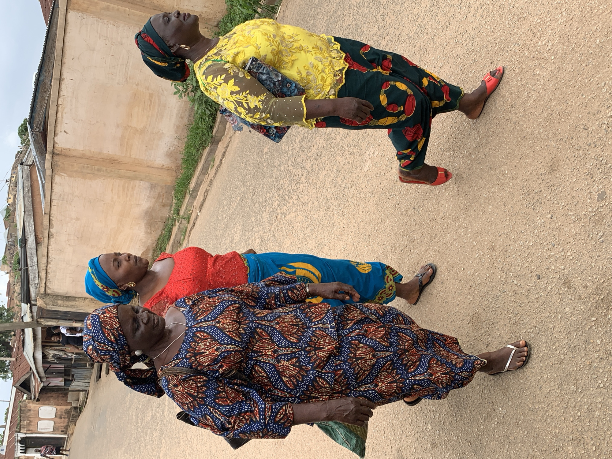 Three African women are walking on a paved road. The woman in the front is wearing a blue and brown patterned top and headwrap. The woman in the middle is wearing a red top and a blue headwrap. The woman in the back is wearing a yellow top and a patterned headwrap. The background consists of buildings and vegetation. Based on the buildings and the style of dress, it is likely the image was taken in Jos, Nigeria.