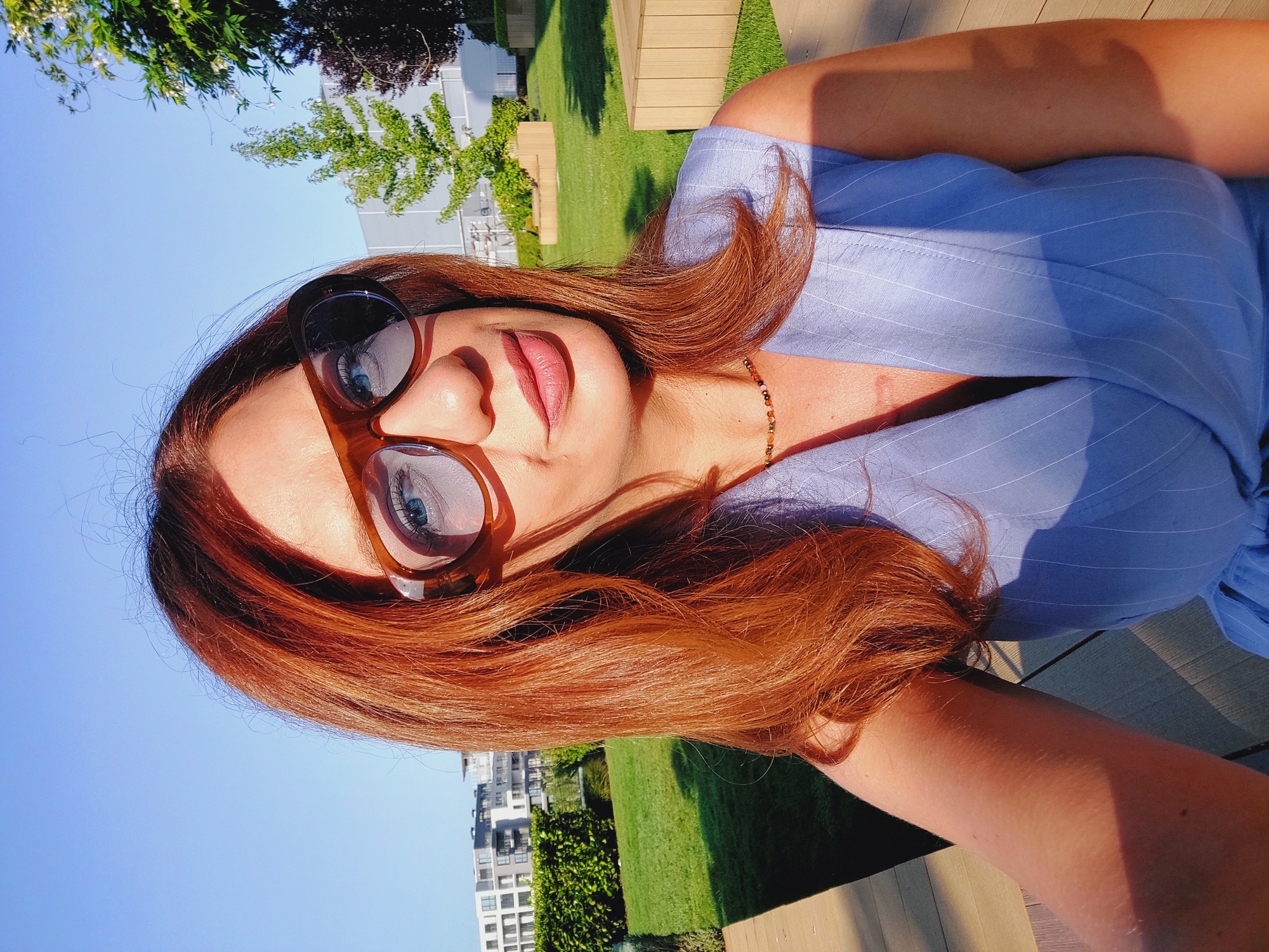 A woman with auburn hair and blue eyes, wearing large brown-rimmed sunglasses and a light blue pinstripe sleeveless dress, is captured in a selfie. She is outdoors in a grassy area with wooden benches and modern buildings in the background. The scene appears to be a park or courtyard, possibly in a contemporary urban development. The lighting suggests it is a sunny day, likely in the late afternoon due to the warm, directional sunlight casting shadows. The woman is looking at the camera, and her expression is relaxed. The backdrop includes lush green grass, manicured hedges, and trees. The buildings in the distance are modern residential or office structures. This location is consistent with new urban developments found in areas such as the Mladost district of Sofia, Bulgaria, which feature modern architecture and green spaces.