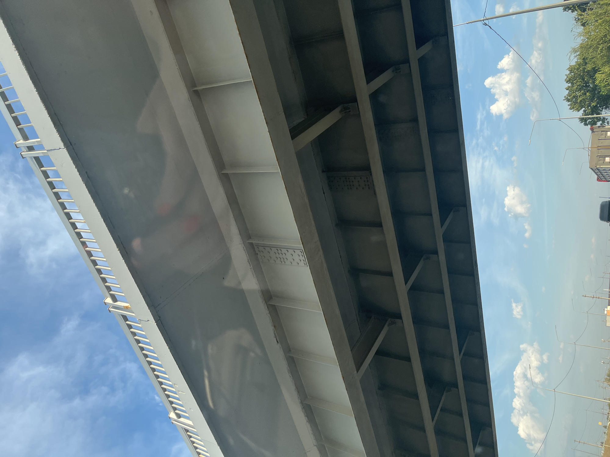 An under-view of a bridge made of concrete and metal beams is visible against a bright blue sky with scattered white clouds. The bridge's underside has a complex structure of reinforced concrete slabs and parallel metal girders, some with visible rivets. Below the bridge, a road can be seen with streetlights, power lines, trees, and a truck. The scene appears to be a roadway under an overpass in the city of Пристанное, Russia.
