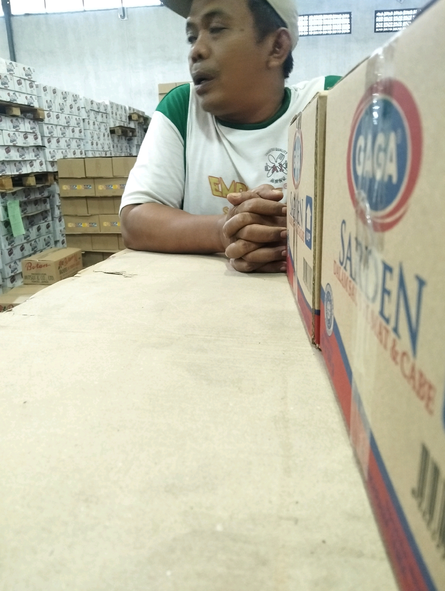 The image depicts a man wearing a white t-shirt with green trim and a light-colored baseball cap, seated in a warehouse environment. He is sitting behind a large cardboard box, with his hands clasped in front of him. The box has prominent branding, including the word "GAGA" in a red and blue circular logo and the word "SARDEN" in large blue letters, along with other text that is partially obscured. In the background, stacks of cardboard boxes are visible, some of which appear to be products on pallets. One visible box in the background has the label "Botan MITSUI & CO., LTD. KOSHIZE, JAPAN". The overall setting suggests a commercial or storage facility. The lighting in the scene is artificial, typical of an indoor warehouse.