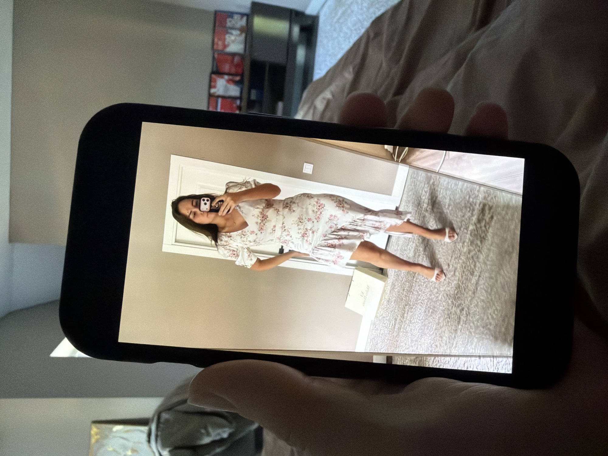 A person is holding a smartphone, displaying a full-body mirror selfie of a woman wearing a white floral dress with pink accents, puffy sleeves, and an asymmetrical ruffled hem. She is also wearing white open-toed heeled sandals. The woman is standing indoors in what appears to be a bedroom, with a bed and a dresser visible in the background. The dresser has several DVDs or Blu-rays stacked on it. A white shopping bag with a logo is on the floor near the woman. The image does not contain any information about Malinovka, Russia.